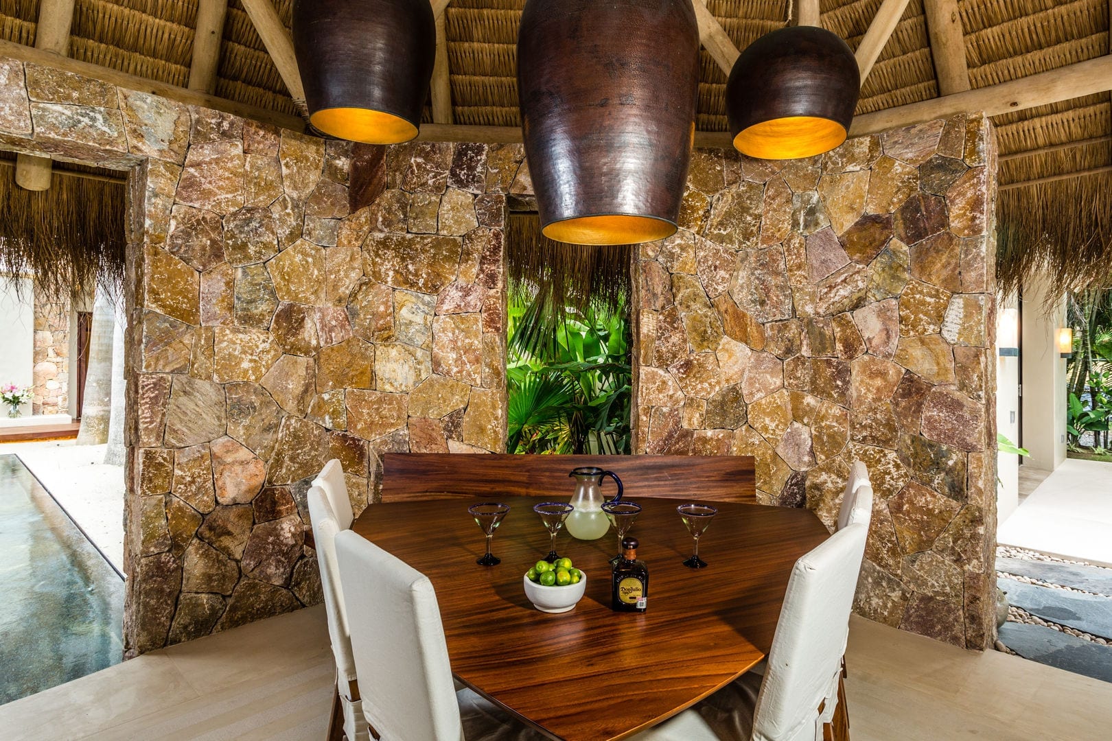 Solid wood dining table set with margarita glasses and fresh limes beneath two large clay pendant lights, rough stone walls and palapa ceiling surrounding the space, Casa Escondida, Ranchos Estates, Punta Mita.