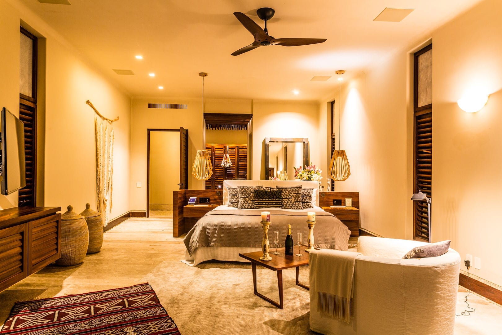 King bed with layered pillows and a low coffee table holding candles and a champagne bottle, carved shuttered windows flanking the room and a woven wall hanging beside the door, Casa Escondida, Ranchos Estates, Punta Mita.