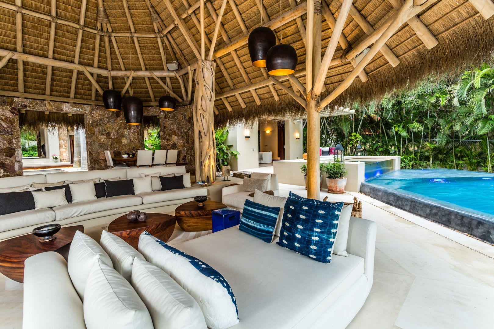 Curved white sectional sofas with indigo-dyed pillows arranged beneath a circular palapa roof, the illuminated pool and jungle visible beyond the open terrace, Casa Escondida, Ranchos Estates, Punta Mita.