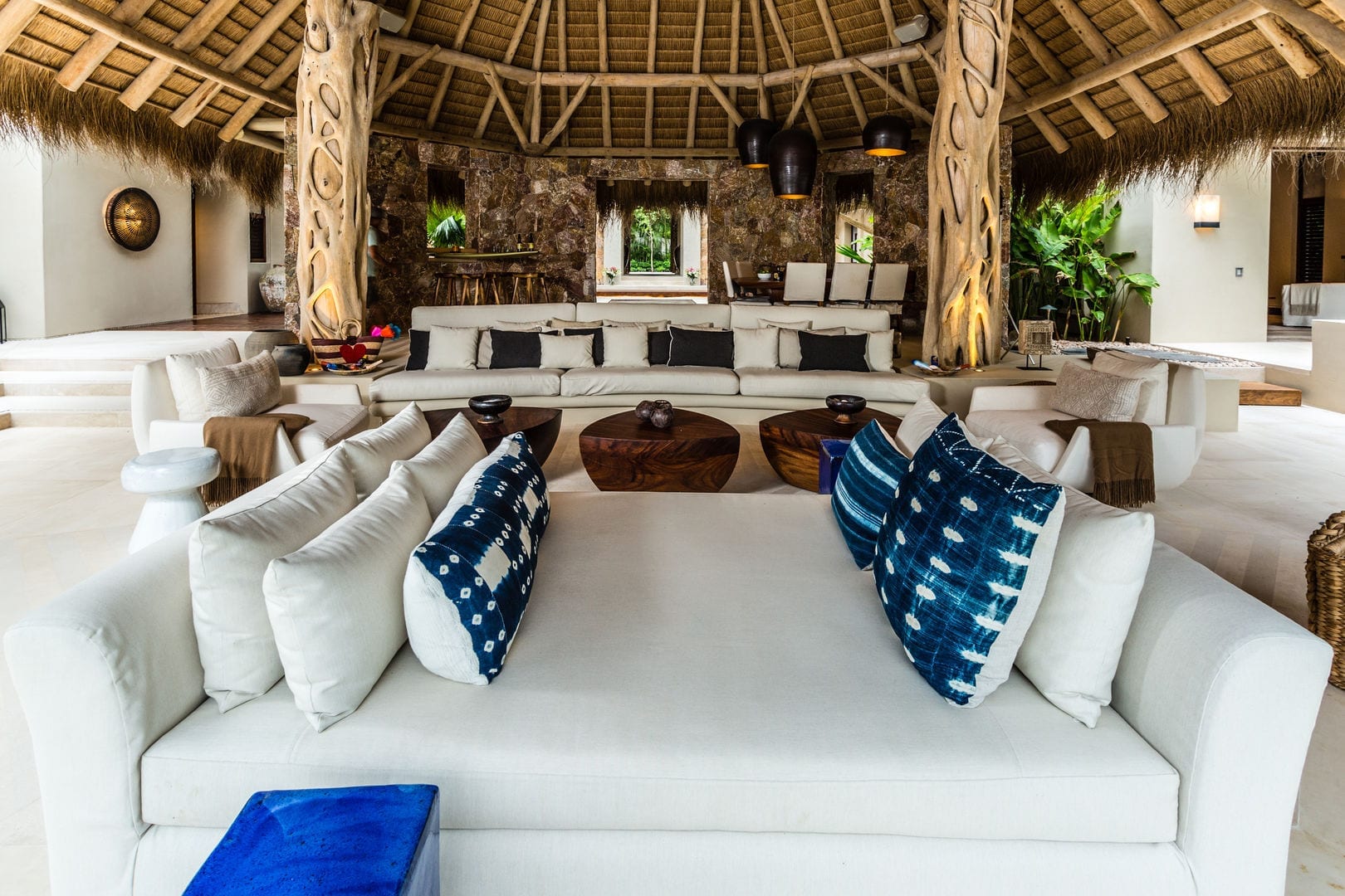 Wide view of the round palapa living pavilion showing carved trunk support columns, a large curved sectional, indigo pillows, wood coffee tables, and the stone bar wall beyond, Casa Escondida, Ranchos Estates, Punta Mita.