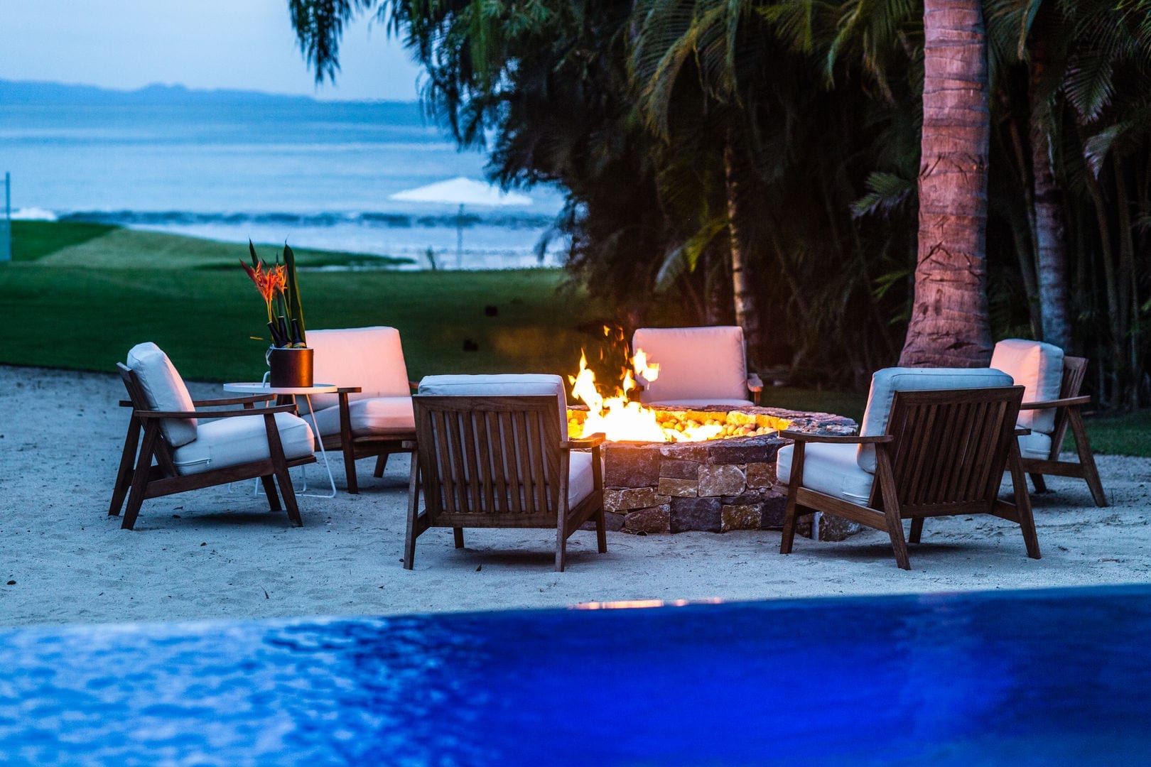 Six teak armchairs arranged around a stone fire pit on the private beach at dusk, the golf fairway and Pacific horizon stretching behind, the pool edge visible in the foreground, Casa Escondida, Ranchos Estates, Punta Mita.