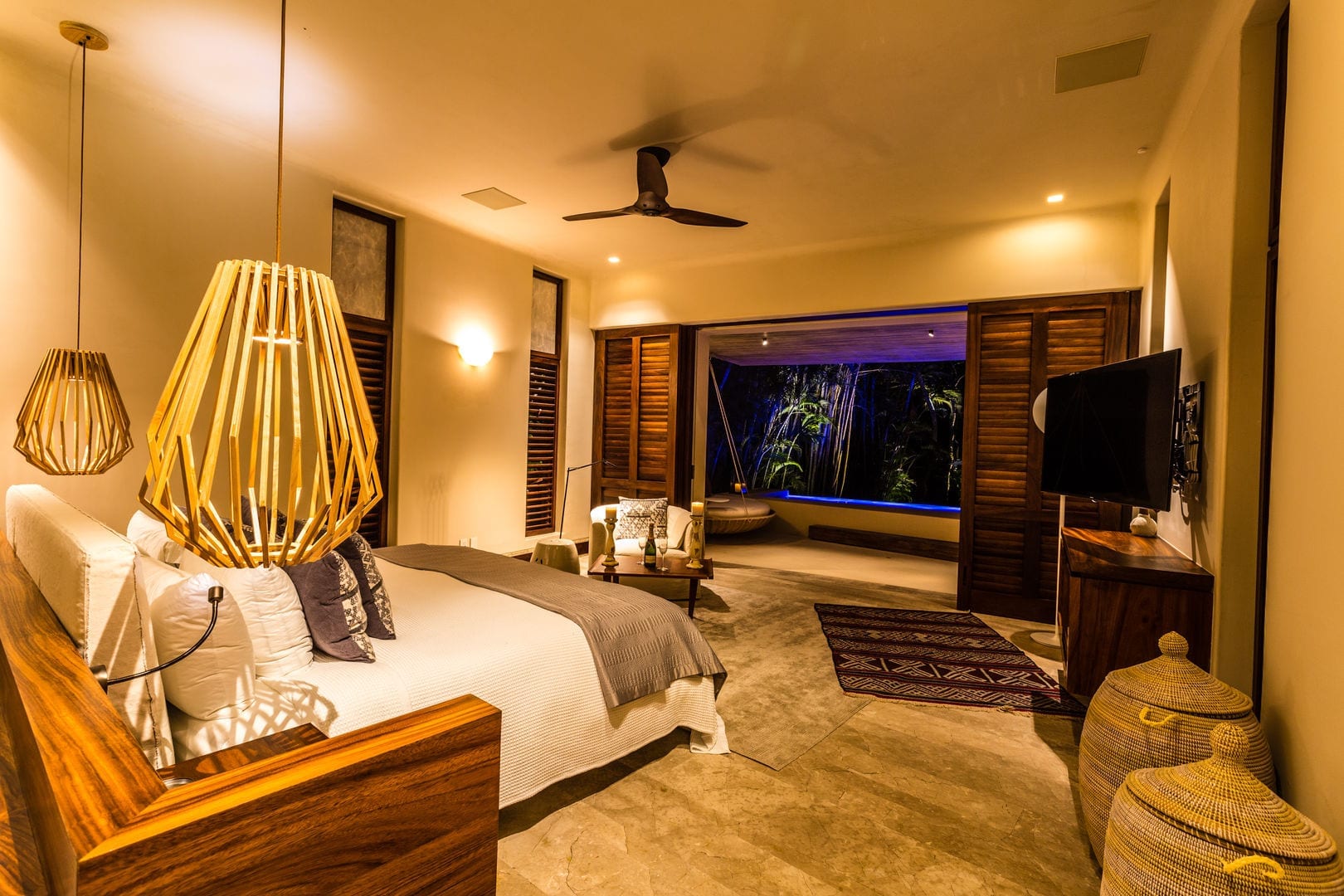 Guest bedroom at night with a dark wood king headboard, geometric rattan pendant light, louvered shutters open to an illuminated garden and pool beyond, wicker baskets at the foot of the bed, Casa Escondida, Ranchos Estates, Punta Mita.