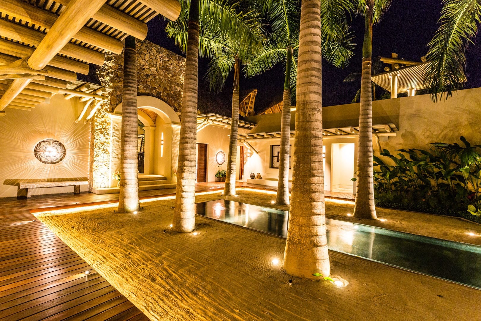 Nighttime view of the courtyard entrance with uplighted palm trunks rising from a hardwood deck, the arched stone entry portal glowing amber ahead, the narrow reflecting pool running along the walkway, Casa Escondida, Ranchos Estates, Punta Mita.