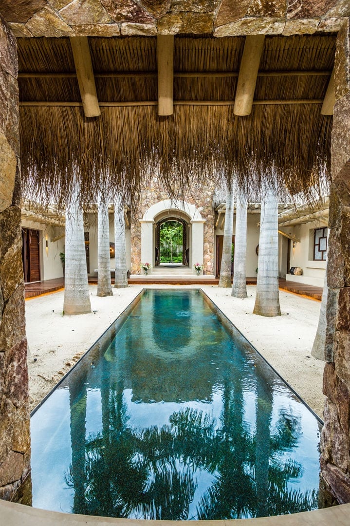 Looking along the courtyard reflecting pool from the palapa end toward the arched stone portal and its garden view beyond, slim palm trunks and rough stone walls framing both sides, Casa Escondida, Ranchos Estates, Punta Mita.