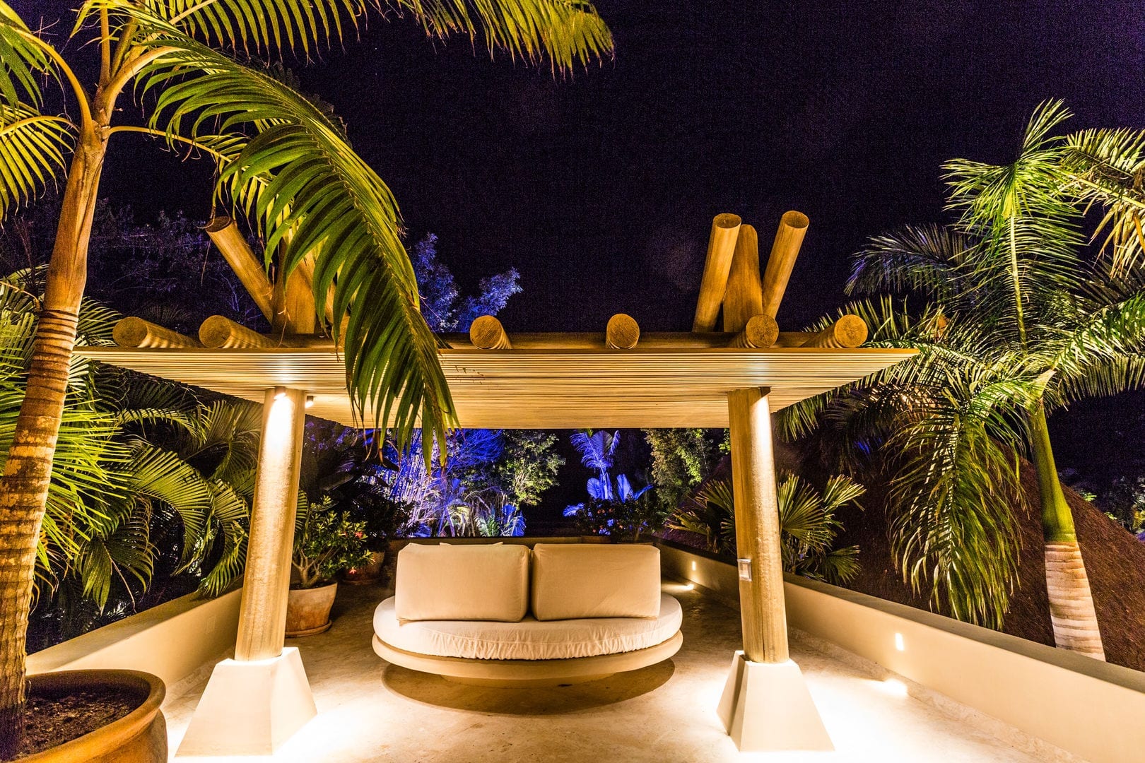 Rooftop terrace at night with a circular cushioned daybed under a low bamboo pergola, uplighted palms surrounding the seating area against a dark sky, Casa Escondida, Ranchos Estates, Punta Mita.