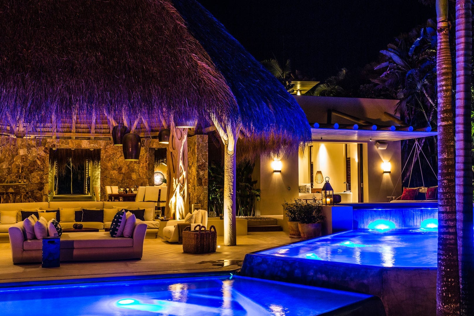 Night view across the pool toward two palapa roofs and the stone-walled living area, warm lamp light spilling from the pavilion while the pool glows blue, Casa Escondida, Ranchos Estates, Punta Mita.