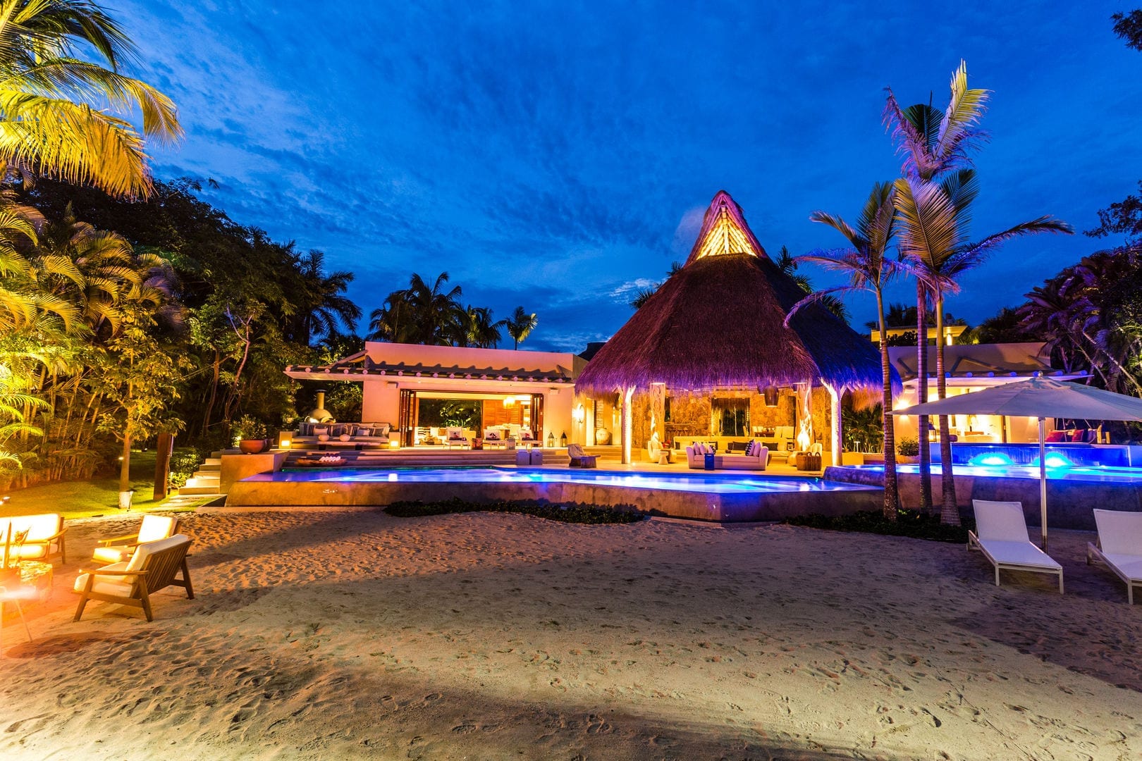 Blue-hour view from the private beach looking toward the palapa pavilion and pool, warm amber lights glowing from inside while the pool shines blue, fire pit chairs on the sand in the foreground, Casa Escondida, Ranchos Estates, Punta Mita.