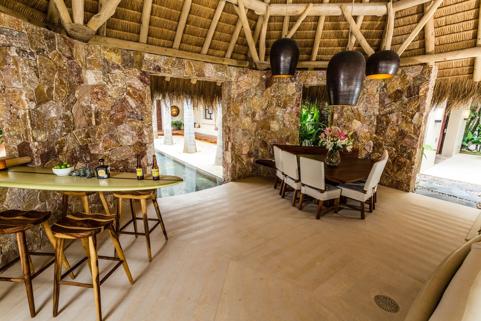 Open-air palapa dining pavilion with rough stone walls, thatched roof, dark gourd pendants, surfboard bar table, and formal dining set for eight, Casa Escondida, Ranchos Estates, Punta Mita.