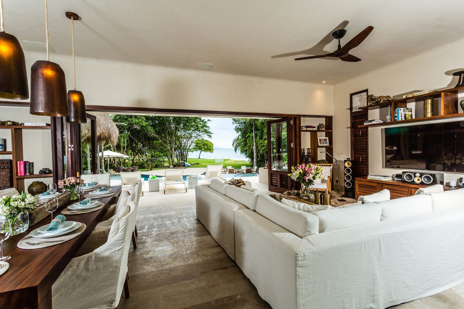 Main house living and dining room with white sectional, dark wood table set for eight, built-in media shelving, and bifold doors open to the pool and ocean, Casa Escondida, Ranchos Estates, Punta Mita.