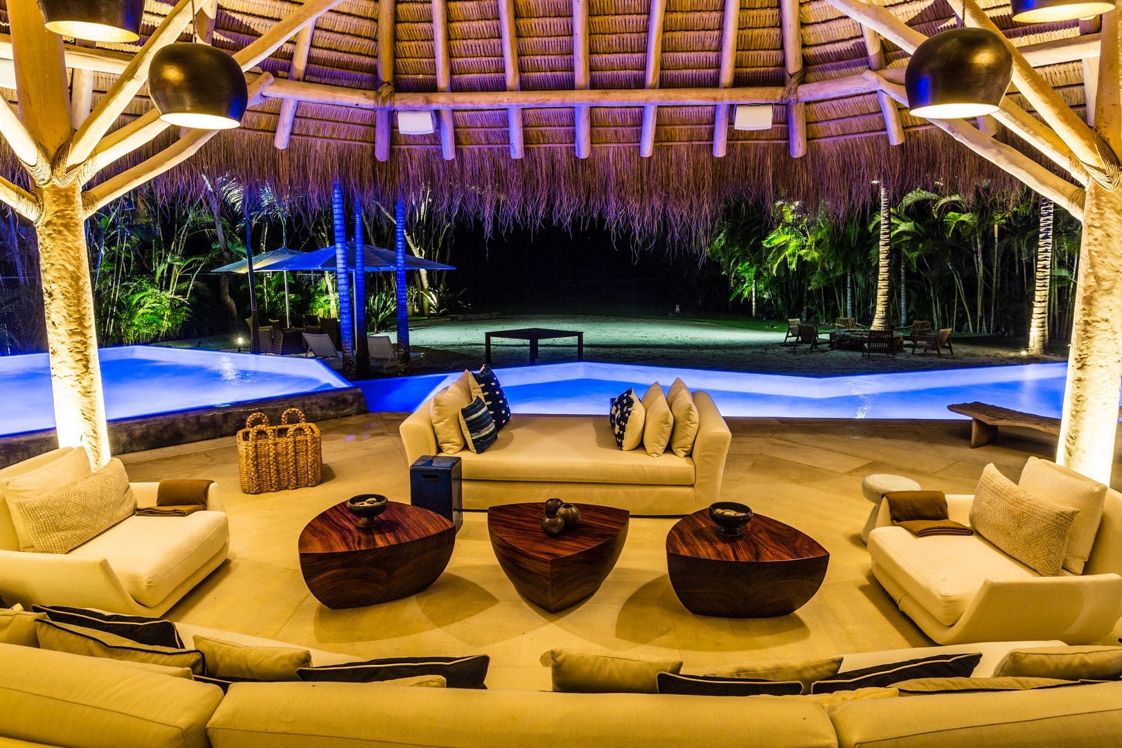 Palapa living pavilion at night with curved sectional sofas, rosewood coffee tables, and the pool glowing blue beyond the open thatch, Casa Escondida, Ranchos Estates, Punta Mita.
