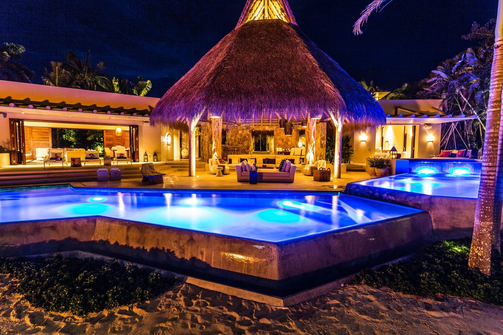Night exterior showing the illuminated pool, palapa pavilion glowing warm at center, main house with open bifold doors at left, and the spa at right, Casa Escondida, Ranchos Estates, Punta Mita.