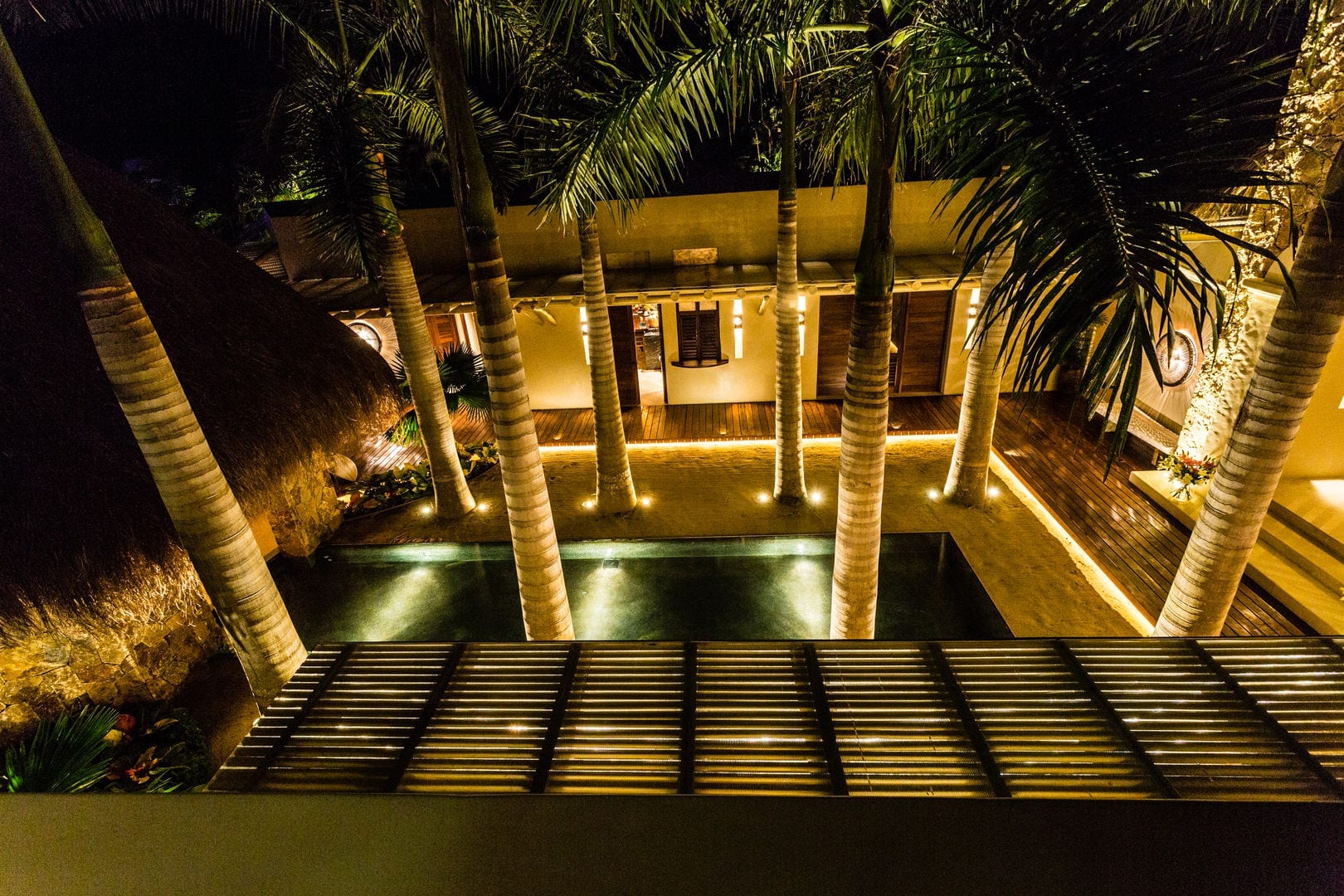 Overhead night view of the long courtyard reflecting pool framed by royal palms with warm uplights on trunks and a slatted deck in the foreground, Casa Escondida, Ranchos Estates, Punta Mita.