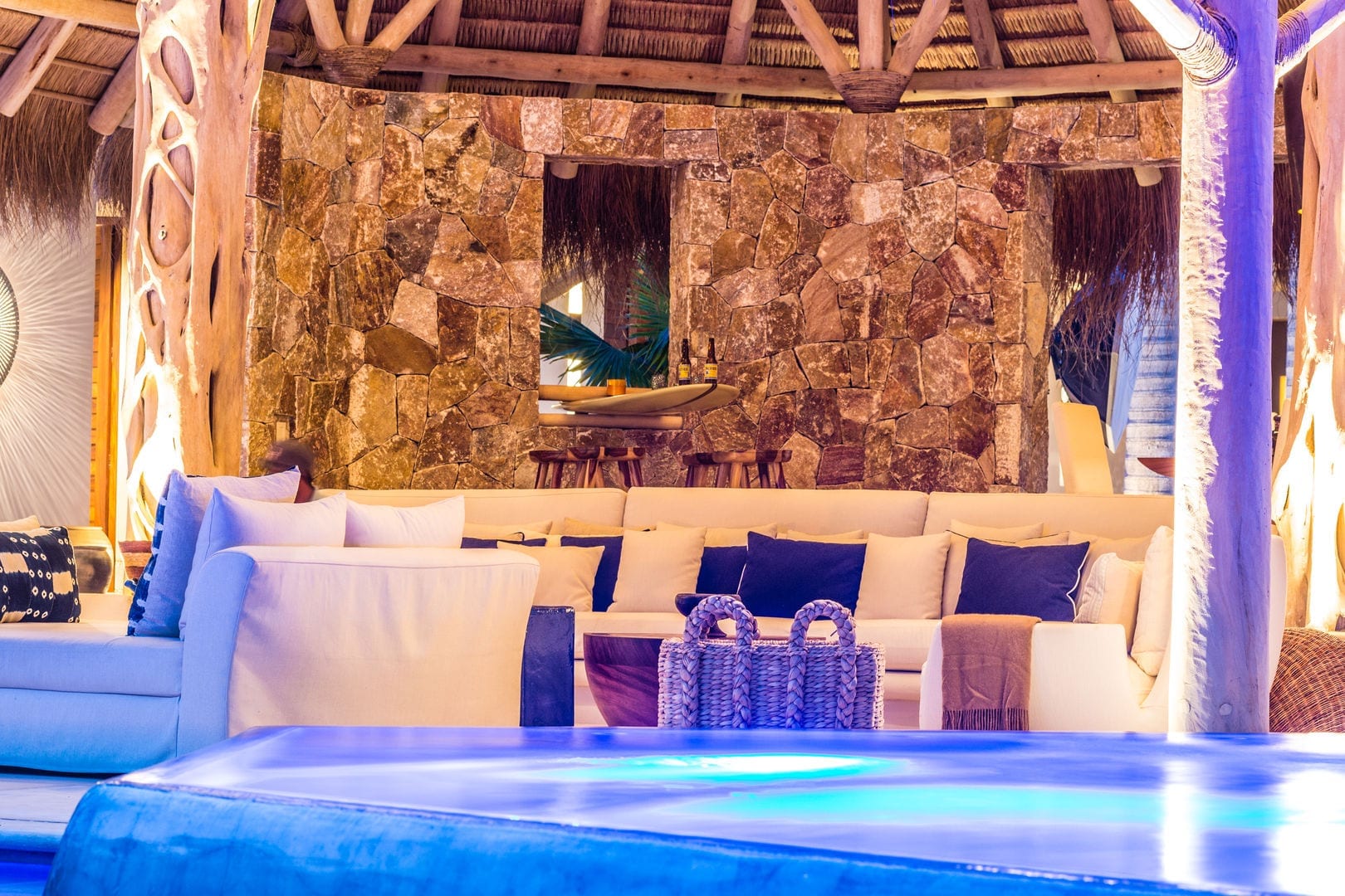 Night view into the palapa pavilion from the pool edge, showing rough stone wall, white sectional seating, and the surfboard bar with stools glowing against the pool's blue light, Casa Escondida, Ranchos Estates, Punta Mita.