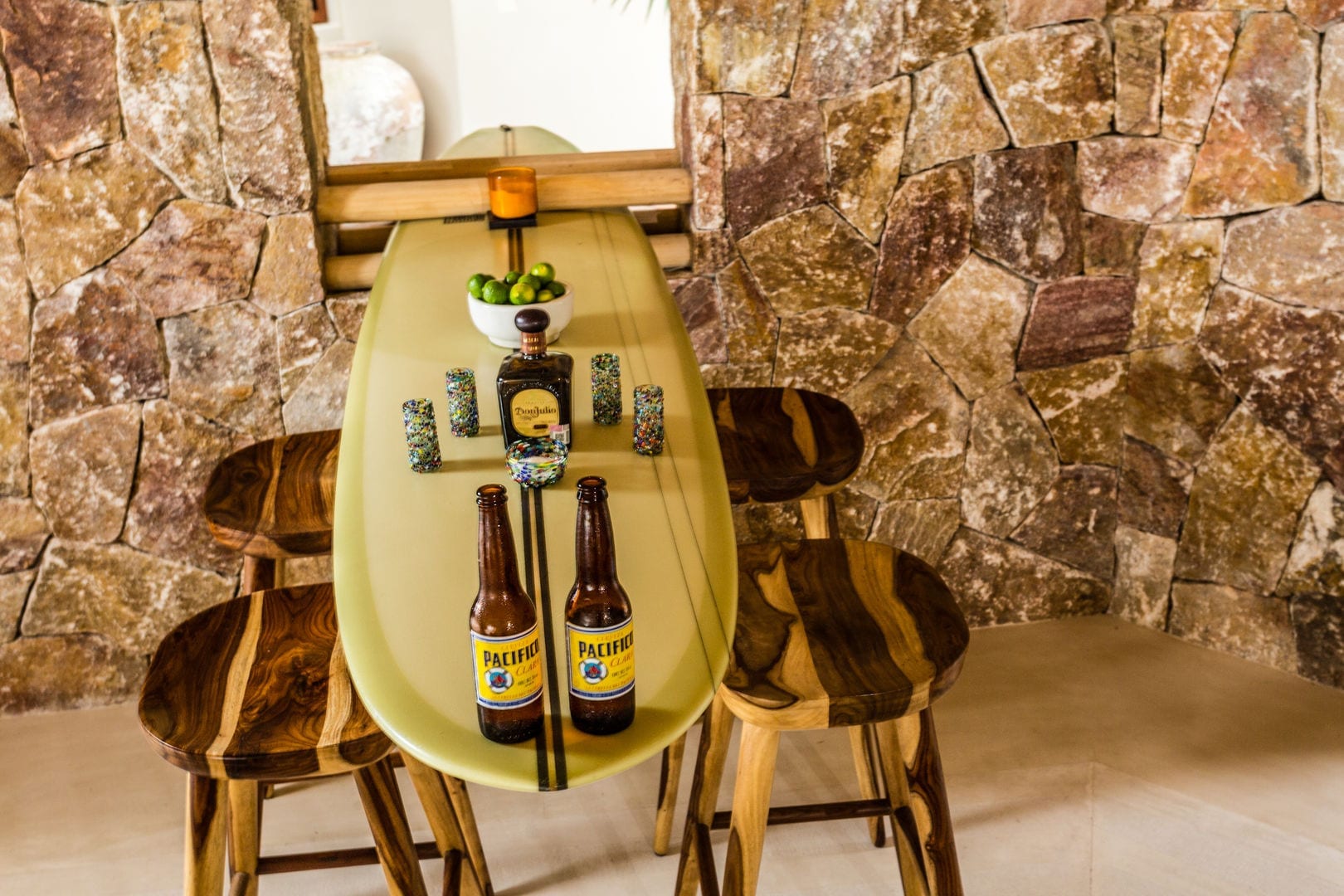 Surfboard bar top set with Don Julio tequila, Pacifico bottles, hand-blown shot glasses, and limes against the rough stone wall of the palapa, Casa Escondida, Ranchos Estates, Punta Mita.