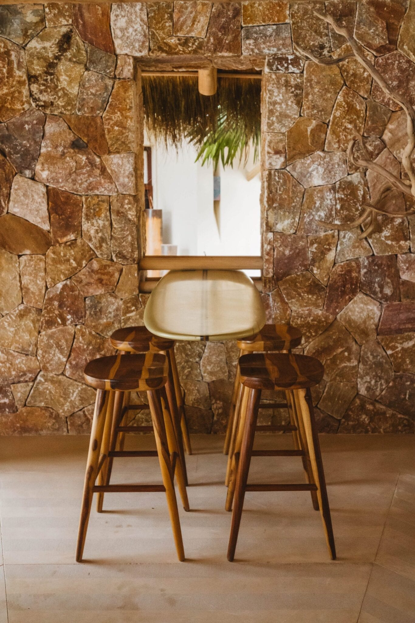 Surfboard bar table with two carved wood stools against the rough stone wall of the palapa, a window frame reflecting thatched fronds and palms beyond, Casa Escondida, Ranchos Estates, Punta Mita.