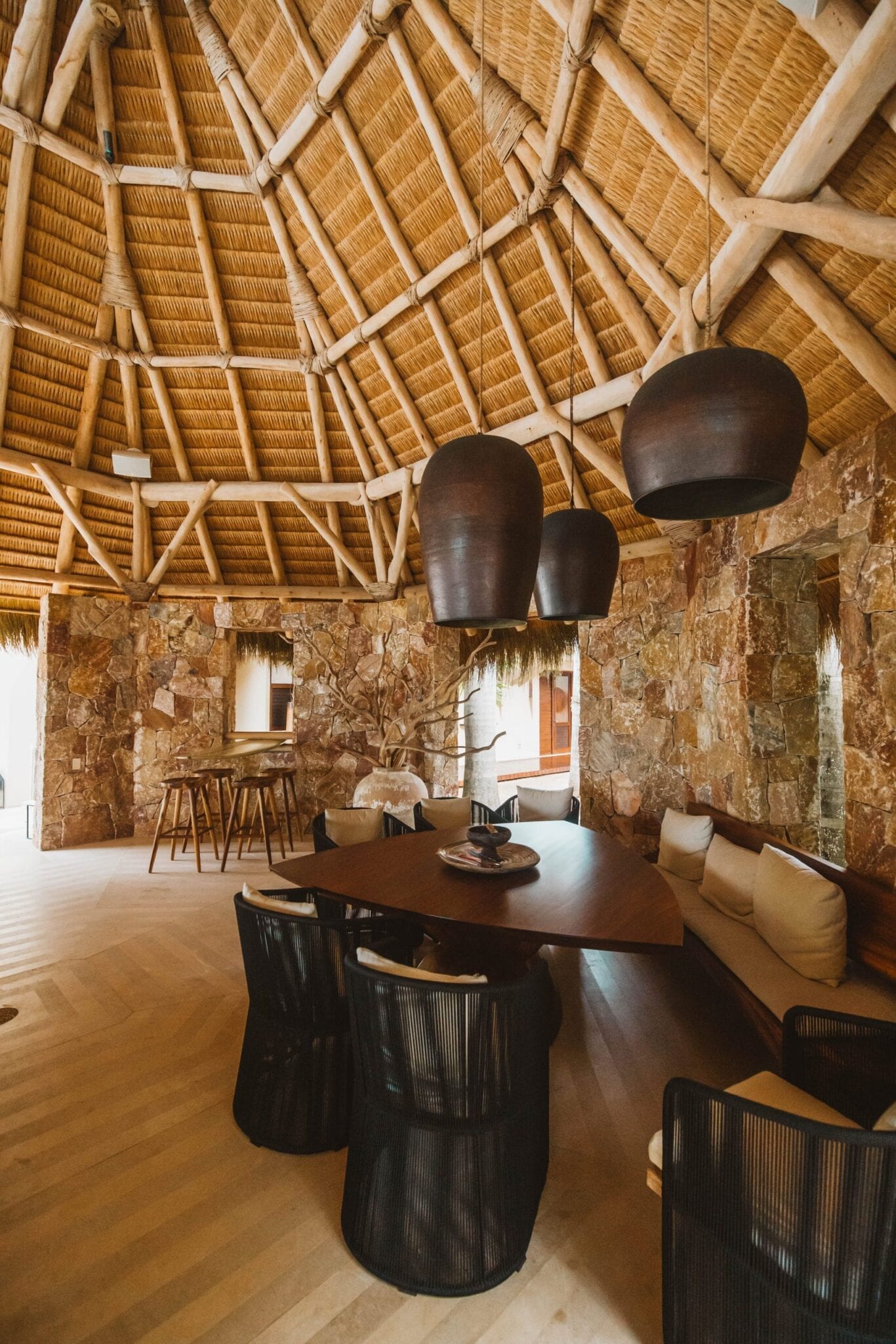 Inside the palapa looking up at the conical thatched roof with log rafters, dark gourd pendants hanging over an oval dining table with woven chairs, and the surfboard bar in the background, Casa Escondida, Ranchos Estates, Punta Mita.