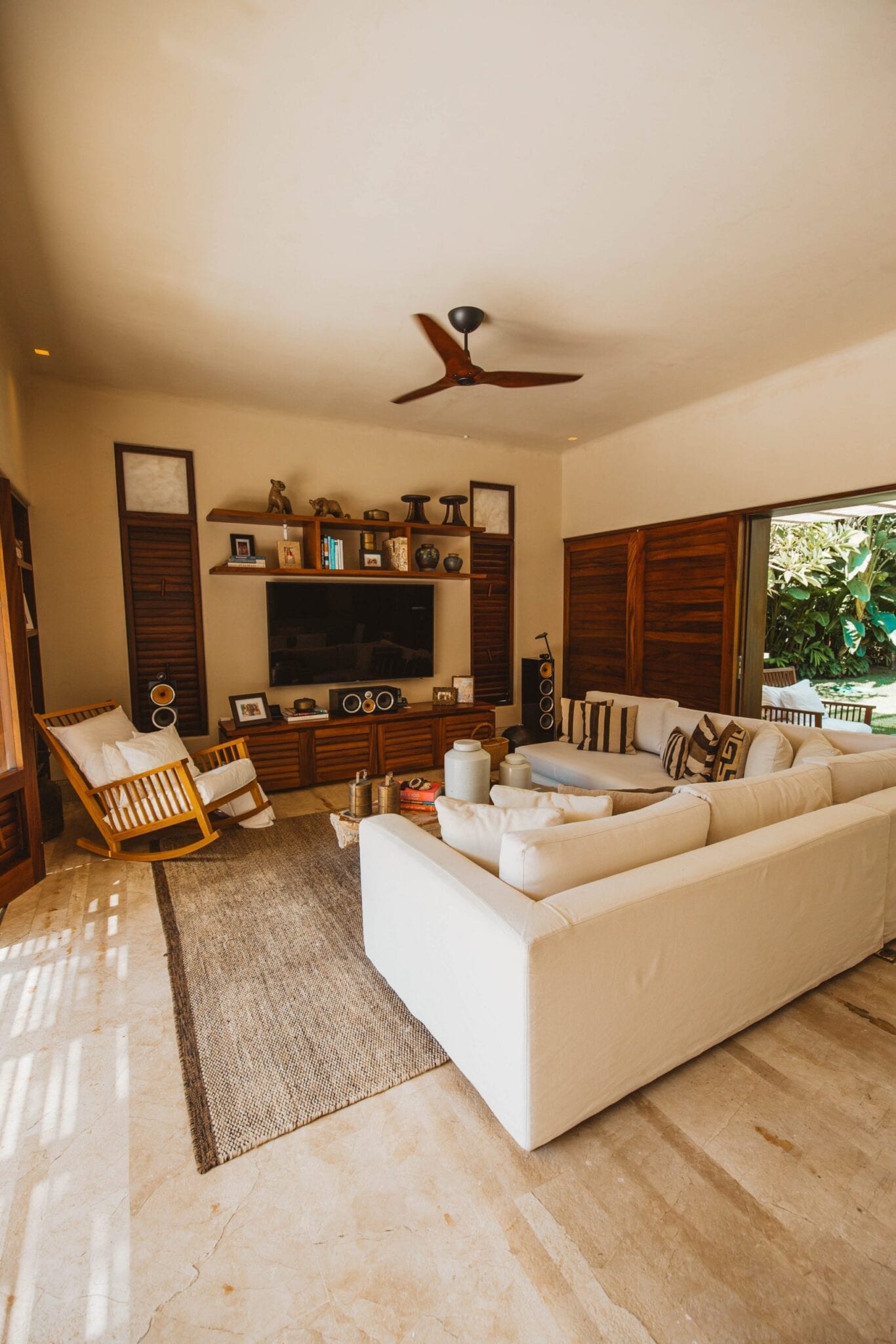 Main house media and living room with white sectional, graphic pillows, flat-screen TV in a built-in wood shelving unit, rocking chair, ceiling fan, and louvered doors open to the garden, Casa Escondida, Ranchos Estates, Punta Mita.