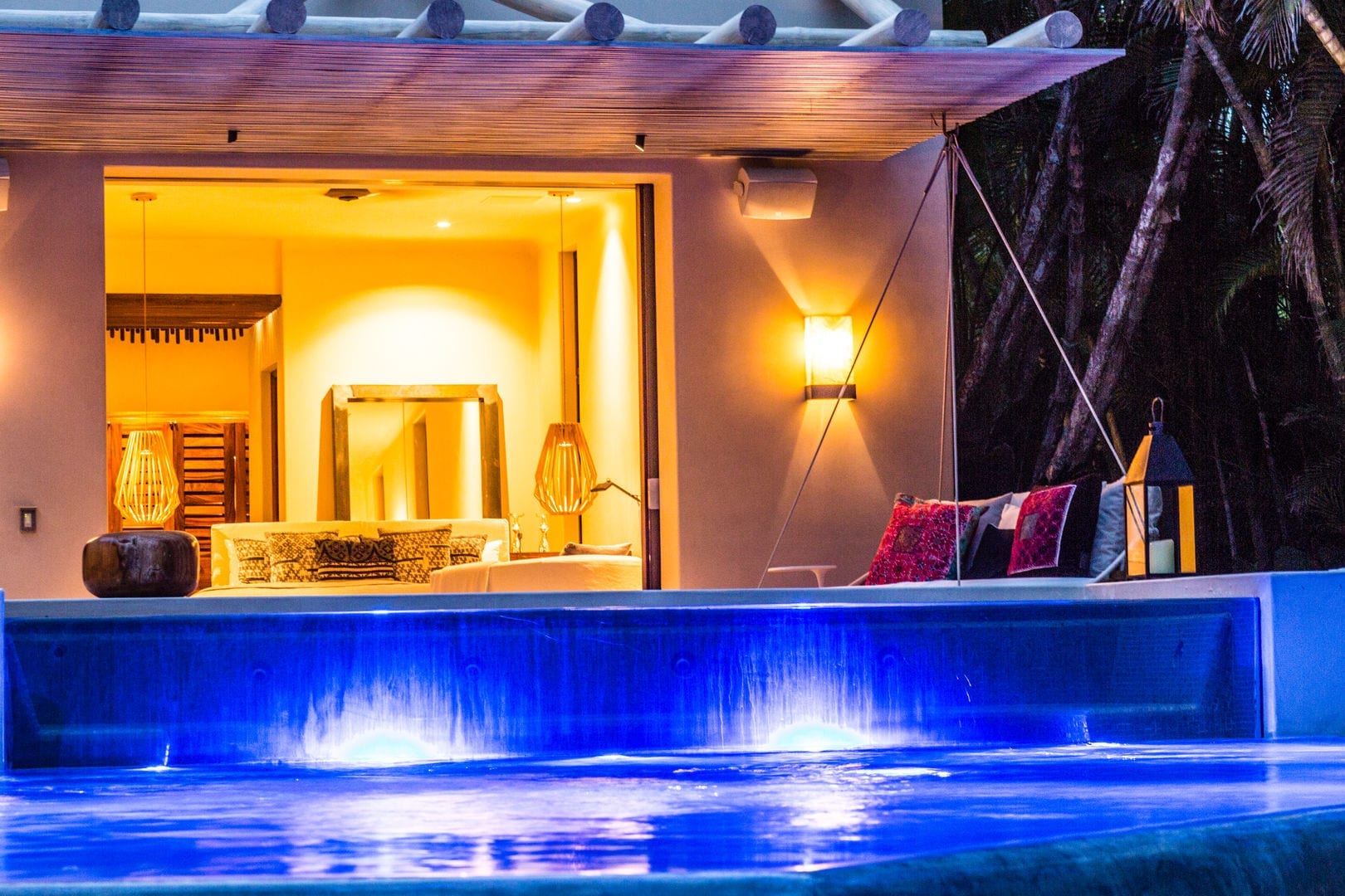 Night view of a bedroom interior glowing warm through fully open sliding glass doors, with a blue-lit pool waterfall cascade in the foreground and an outdoor terrace with lantern and cushioned seating, Casa Escondida, Ranchos Estates, Punta Mita.