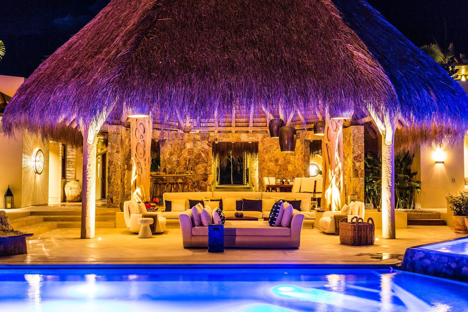 Night exterior of the palapa pavilion lit in warm amber with stone walls, tree-trunk columns, and sofas visible inside, the pool glowing blue in the foreground, Casa Escondida, Ranchos Estates, Punta Mita.