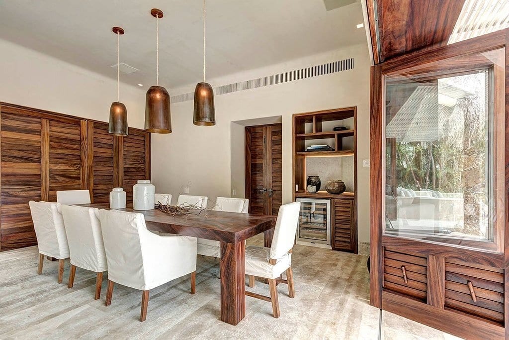 Indoor formal dining room with a long dark wood table, eight white slipcovered chairs, three copper pendants, wood-paneled walls, built-in bar niche with wine fridge, and glass doors to the garden, Casa Escondida, Ranchos Estates, Punta Mita.