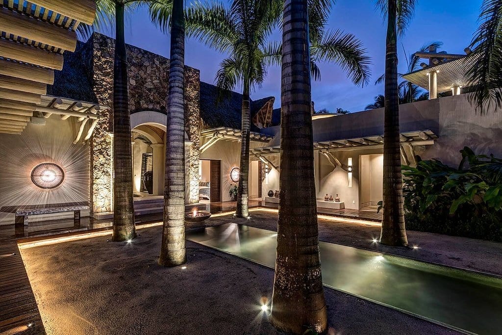 Twilight view of the arched stone entry courtyard with long reflecting pool channels, royal palms lit from below, and the palapa roof edge at left, Casa Escondida, Ranchos Estates, Punta Mita.