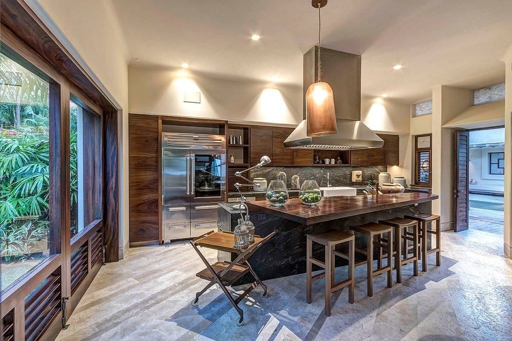 Kitchen with dark granite island, copper pendant, stainless range hood, Sub-Zero refrigerator, dark wood cabinetry, limestone tile floor, bar stools, and a garden-view window, Casa Escondida, Ranchos Estates, Punta Mita.