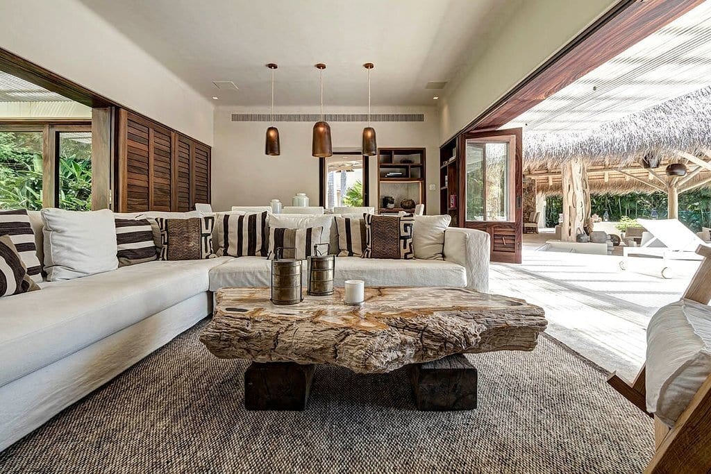 Main house living room with white sectional, graphic throw pillows, a large petrified-wood coffee table, copper pendants, and open bifold doors to the pool terrace and palapa beyond, Casa Escondida, Ranchos Estates, Punta Mita.