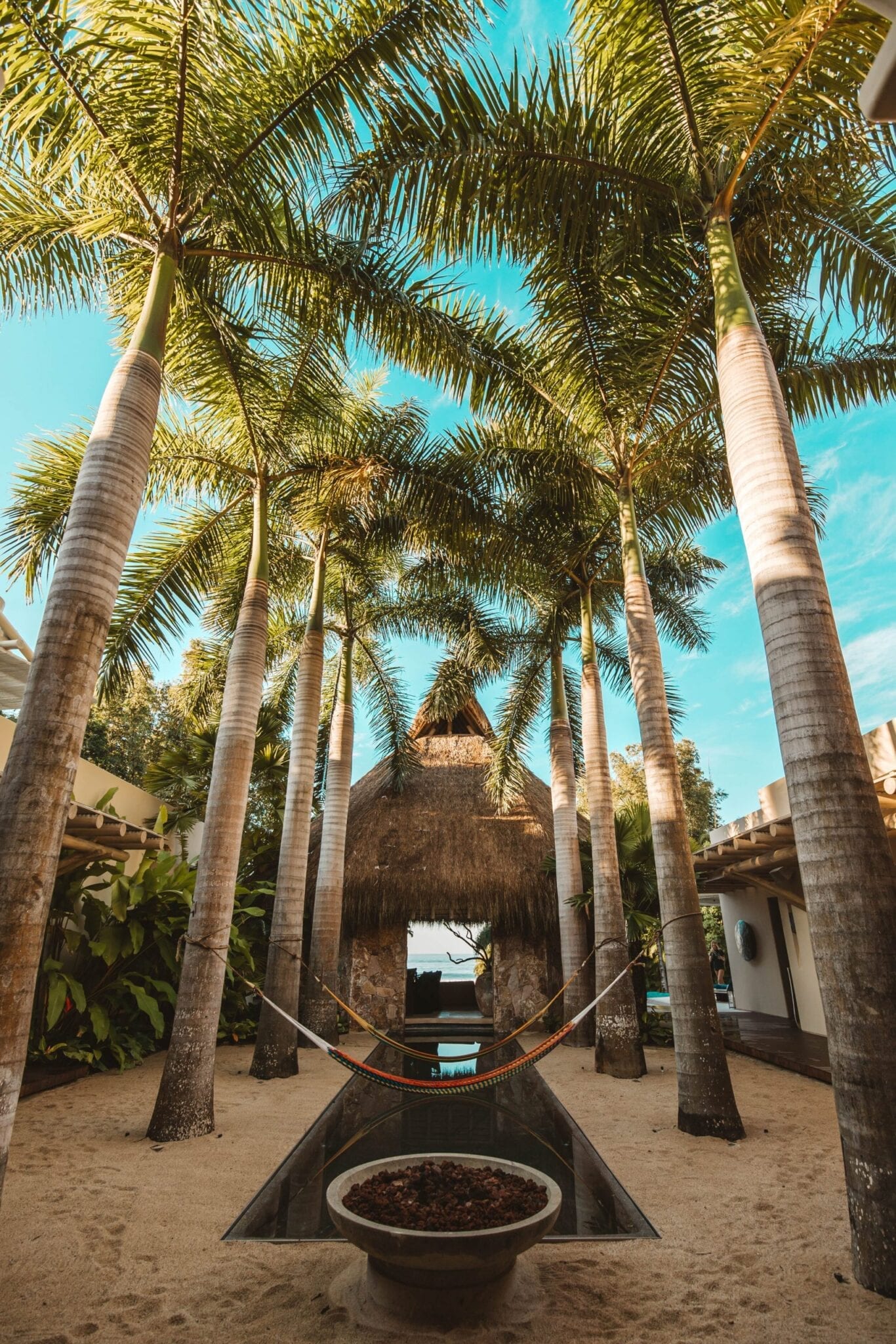 Looking up through a colonnade of royal palms toward the palapa pavilion, with a hammock strung between trunks, a fire bowl in the foreground, and the pool beyond, Casa Escondida, Ranchos Estates, Punta Mita.