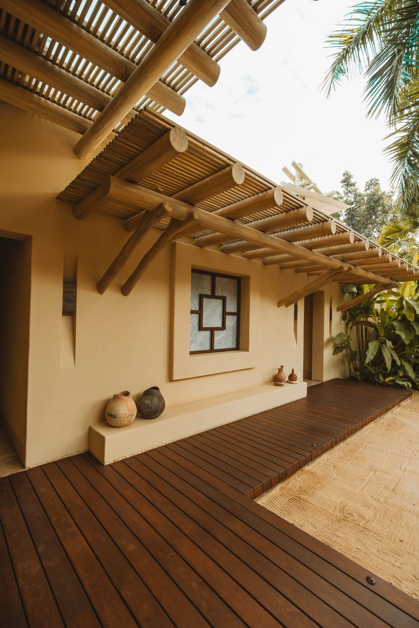 Exterior walkway with a slatted log pergola overhead, cream stucco wall, decorative clay pots on a low bench, and a hardwood deck bordered by tropical foliage, Casa Escondida, Ranchos Estates, Punta Mita.