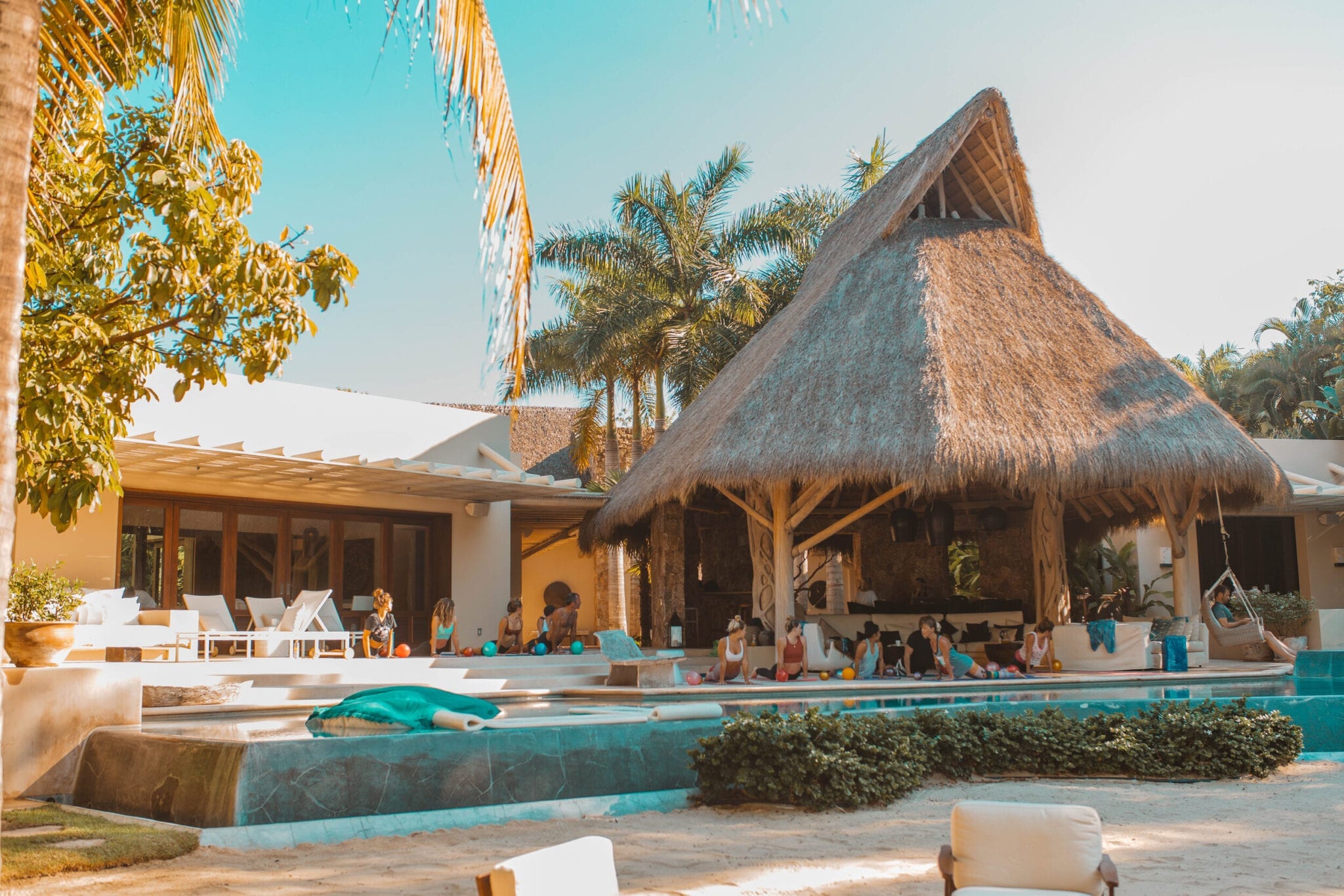 Pool terrace with guests gathered under the palapa pavilion, lounge chairs along the pool edge, the main house pergola behind, and palm trees framing a blue sky, Casa Escondida, Ranchos Estates, Punta Mita.
