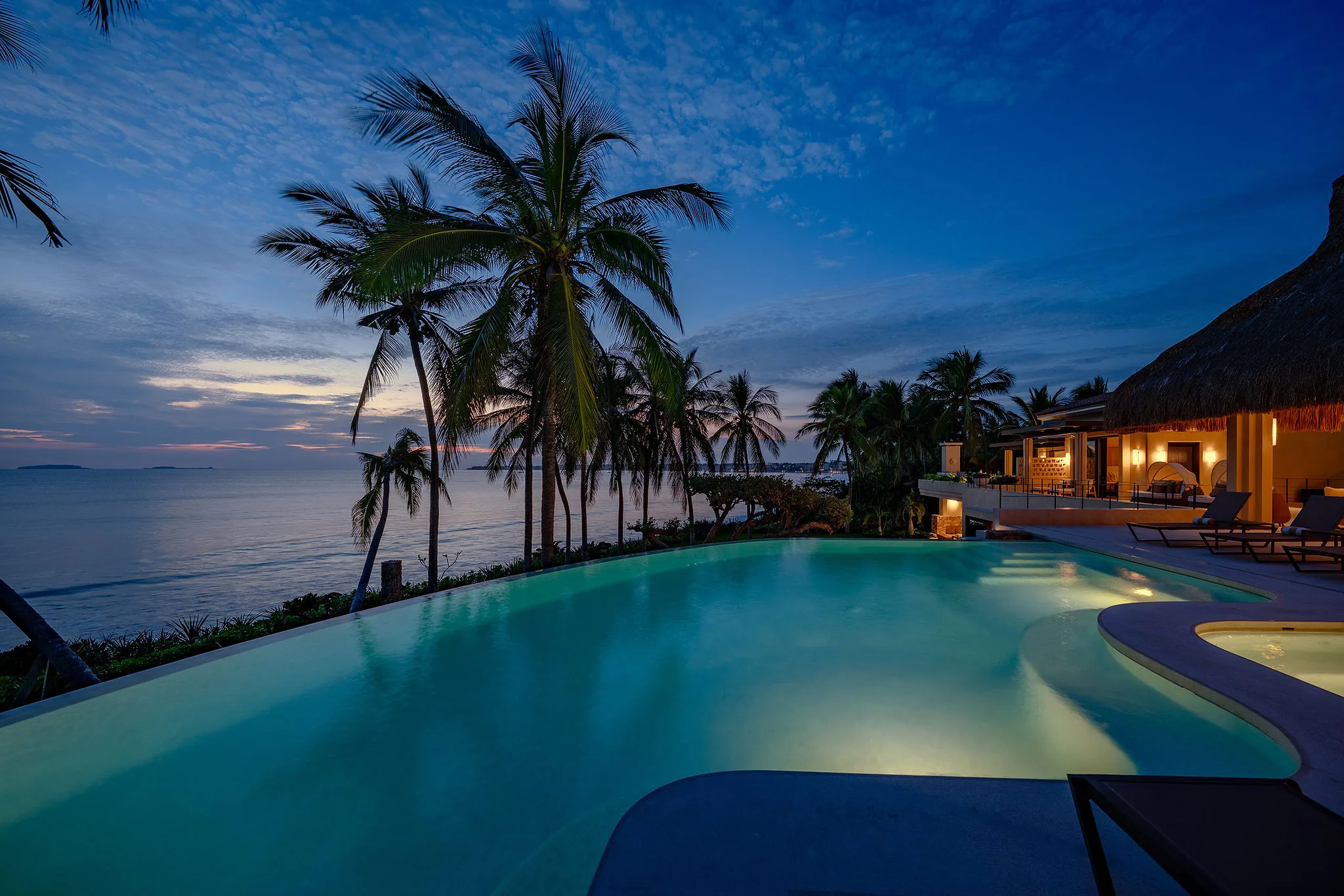 Infinity pool glowing turquoise at blue hour, coconut palms silhouetted against the open ocean, thatched bar and living pavilion lit to the right, Casa Happy Paws, Ranchos Estates, Punta Mita.