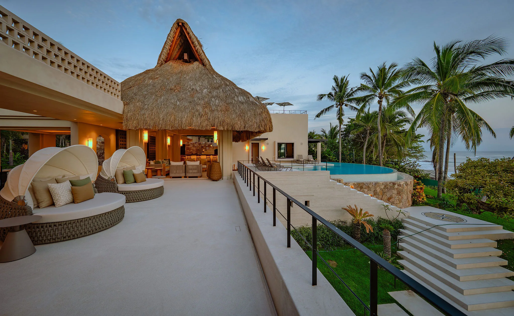 Upper terrace at dusk showing two round canopied daybeds, the thatched palapa pavilion lit warmly, the crescent pool with white curved coping, and the Pacific beyond, Casa Happy Paws, Ranchos Estates, Punta Mita.