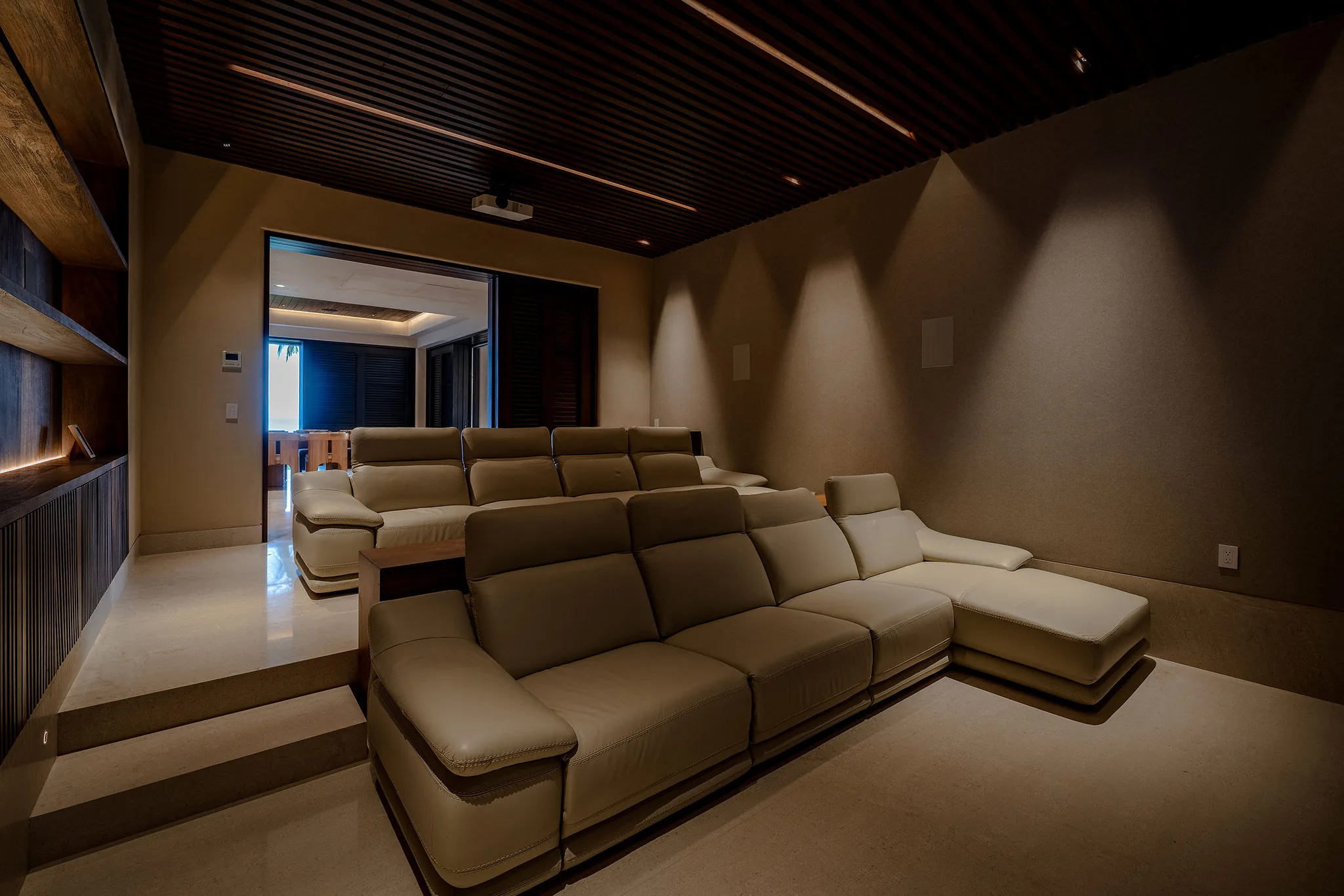 Private home theater with two tiered rows of cream leather reclining seats, a ceiling-mounted projector, slat-wood ceiling with amber cove lighting, and a hallway opening to the villa beyond, Casa Happy Paws, Ranchos Estates, Punta Mita.