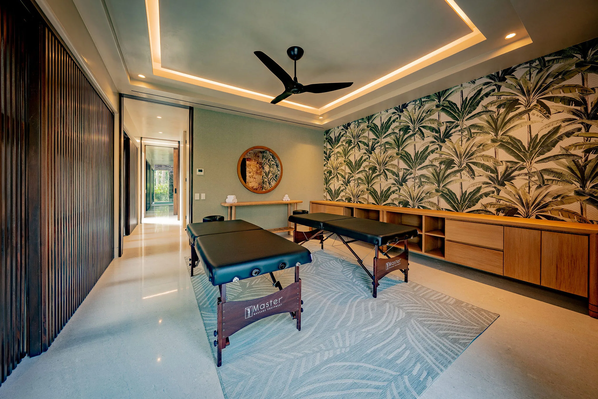 Spa treatment room with two portable massage tables on a patterned aqua rug, a tropical-palm wallpaper accent wall, tray ceiling with cove lighting, and ceiling fan overhead, Casa Happy Paws, Ranchos Estates, Punta Mita.