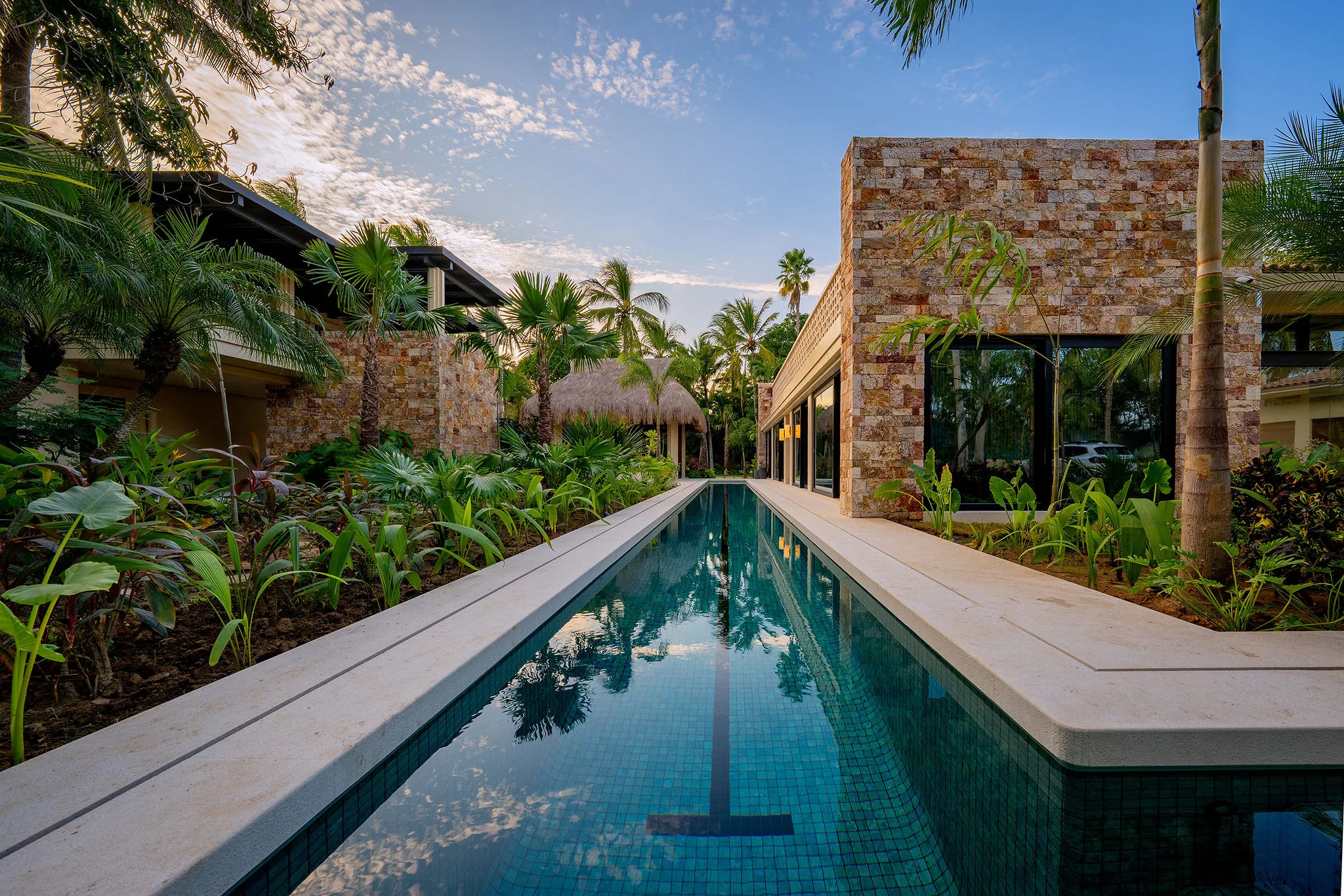 Narrow lap pool reflecting the sky, flanked by dense tropical plantings and textured stone walls, with a small thatched structure visible among the palms at the far end, Casa Happy Paws, Ranchos Estates, Punta Mita.
