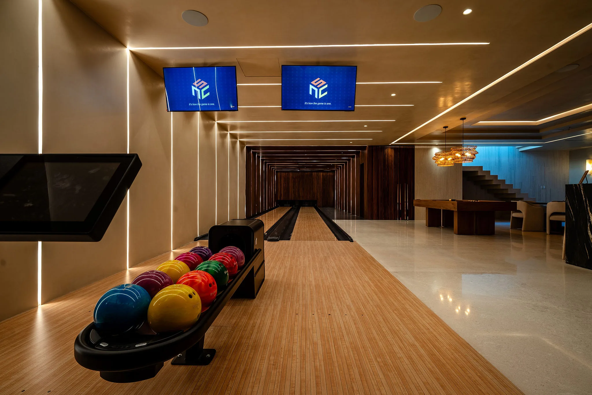 Two-lane private bowling alley with a rack of colorful balls in the foreground, dual scoring monitors overhead, linear LED ceiling strips, shuffleboard table and lounge seating visible in the adjacent game room, Casa Happy Paws, Ranchos Estates, Punta Mita.