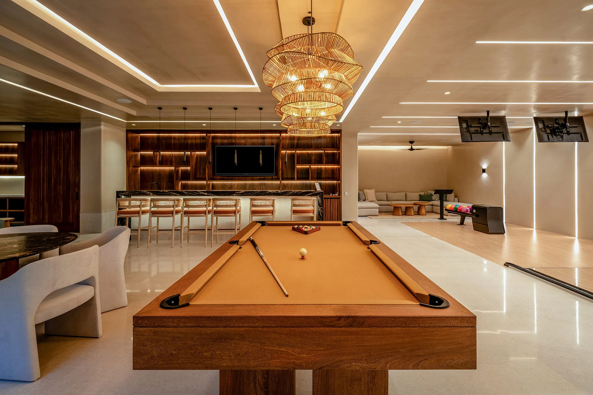 Billiards table under a tiered woven rattan chandelier, with a full marble-top bar and bar stools in the background, TV wall unit flanked by dark wood shelving, and the bowling lane lit beyond, Casa Happy Paws, Ranchos Estates, Punta Mita.