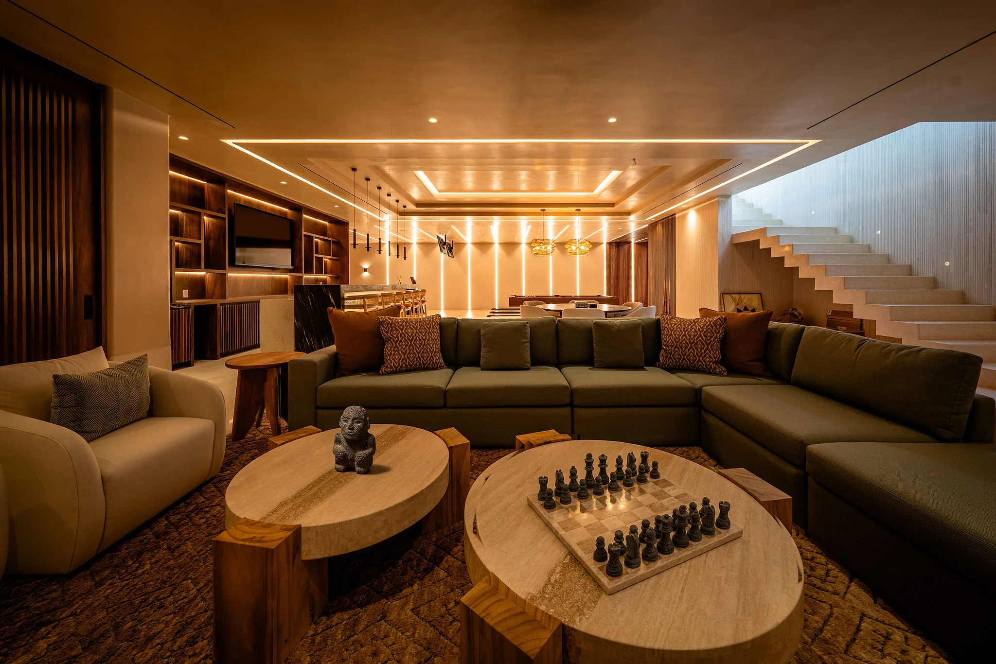 Lounge seating area of the game room with a chess set and pre-Columbian figurine on paired travertine coffee tables, a large olive green sectional sofa, backlit shelving and bar unit in the background, Casa Happy Paws, Ranchos Estates, Punta Mita.