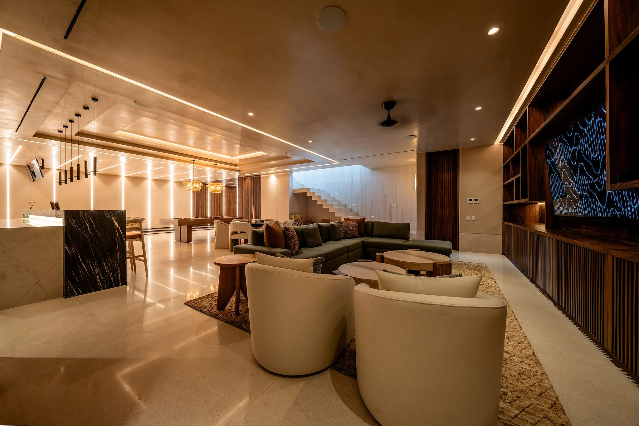 Game room from the TV-wall end showing two curved cream armchairs in the foreground, green sectional sofa behind, the black marble bar extending right, and the bowling lane entrance glowing at far left, Casa Happy Paws, Ranchos Estates, Punta Mita.
