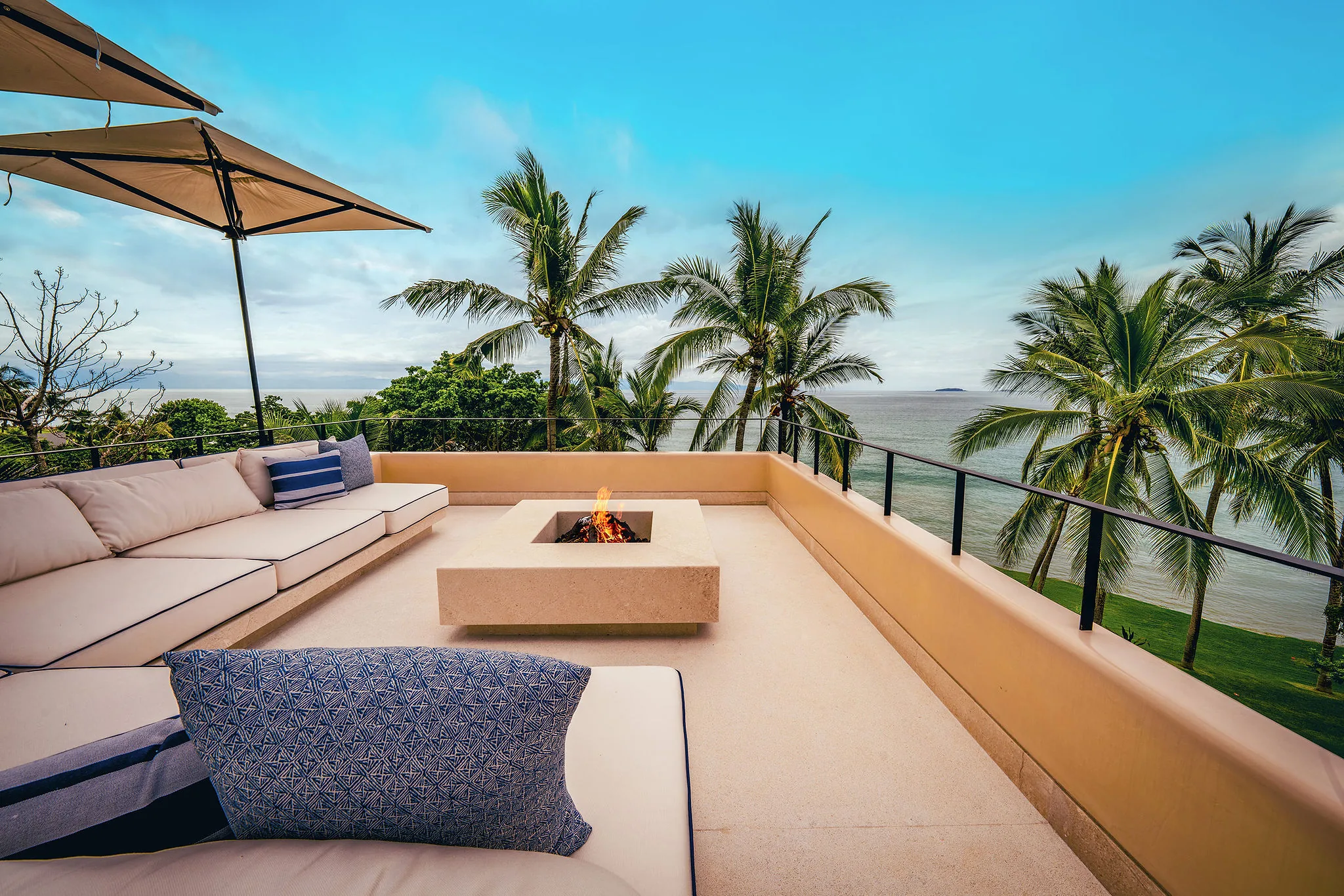 Rooftop sky terrace with a square concrete fire pit burning, built-in white bench sofas, a market umbrella, cable-rail perimeter, palms and the Pacific with distant islands framed beyond, Casa Happy Paws, Ranchos Estates, Punta Mita.