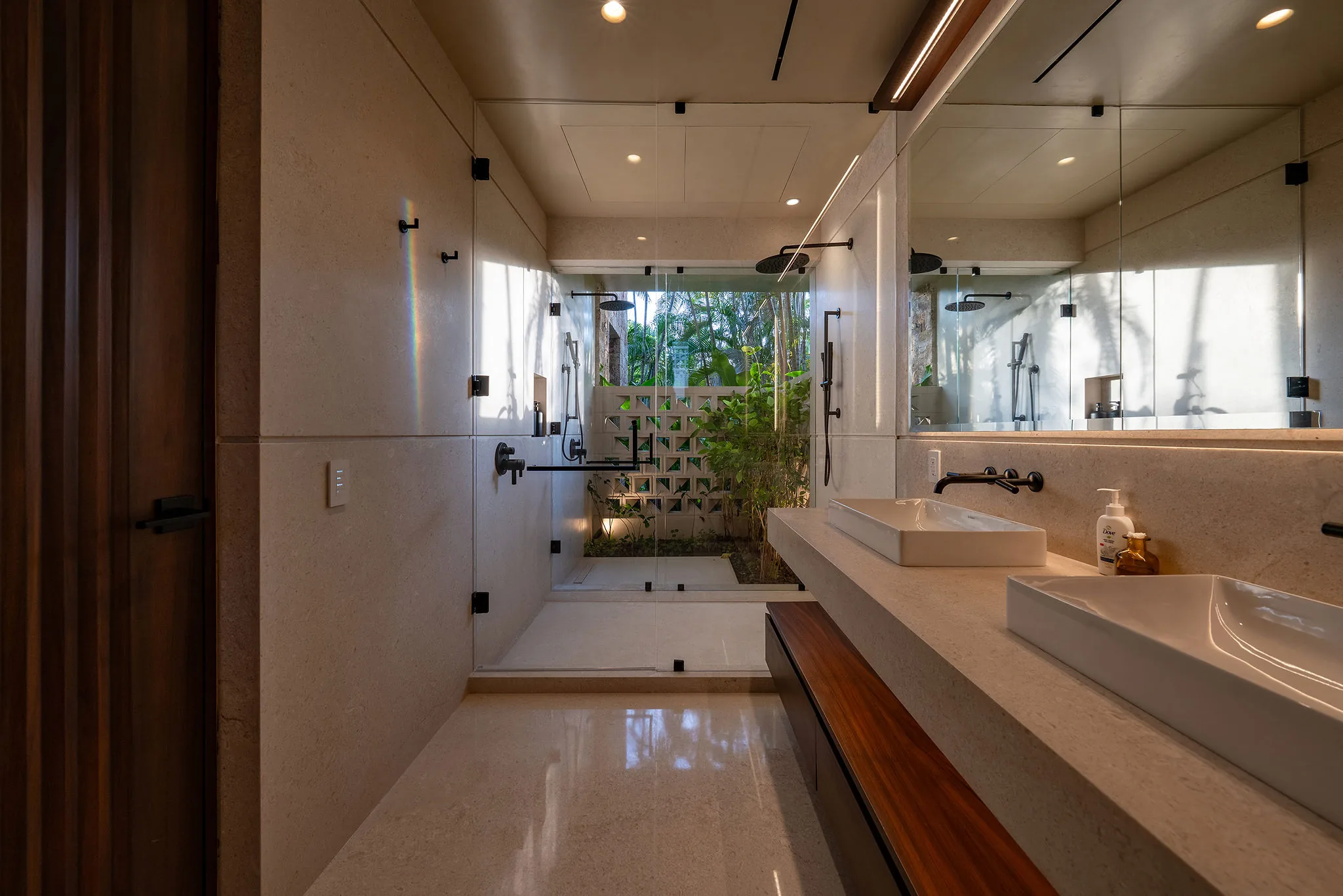 Ensuite bath with double vessel sinks on a wood-and-stone vanity, wide mirror, and glass shower opening to a lattice garden wall, Casa Happy Paws, Ranchos Estates, Punta Mita.