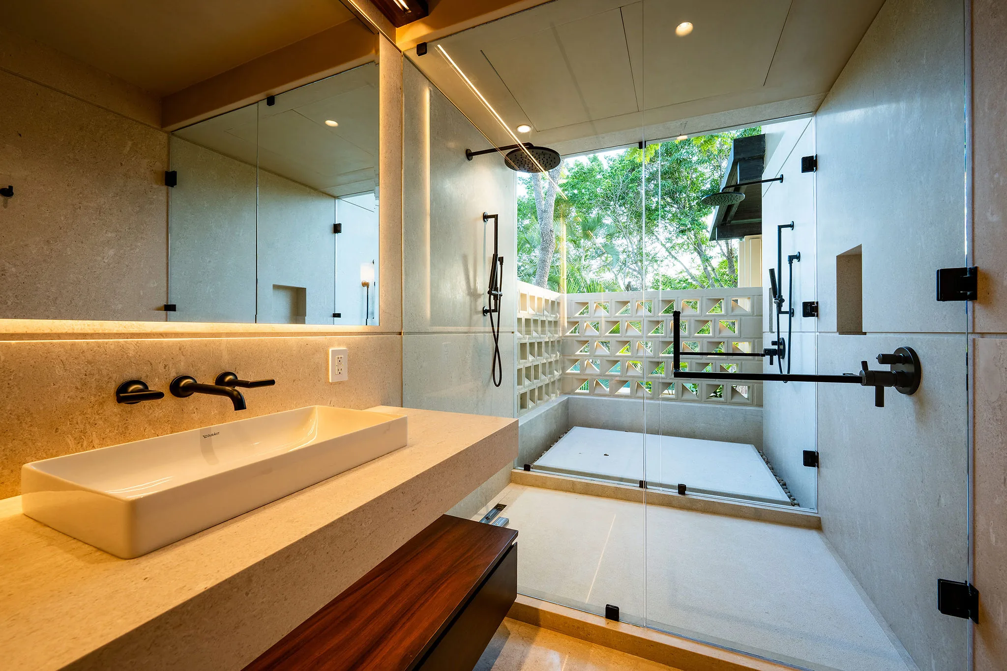 Ensuite bath with single vessel sink on stone-and-teak vanity, rain-head glass shower, and open lattice garden wall with tree canopy above, Casa Happy Paws, Ranchos Estates, Punta Mita.