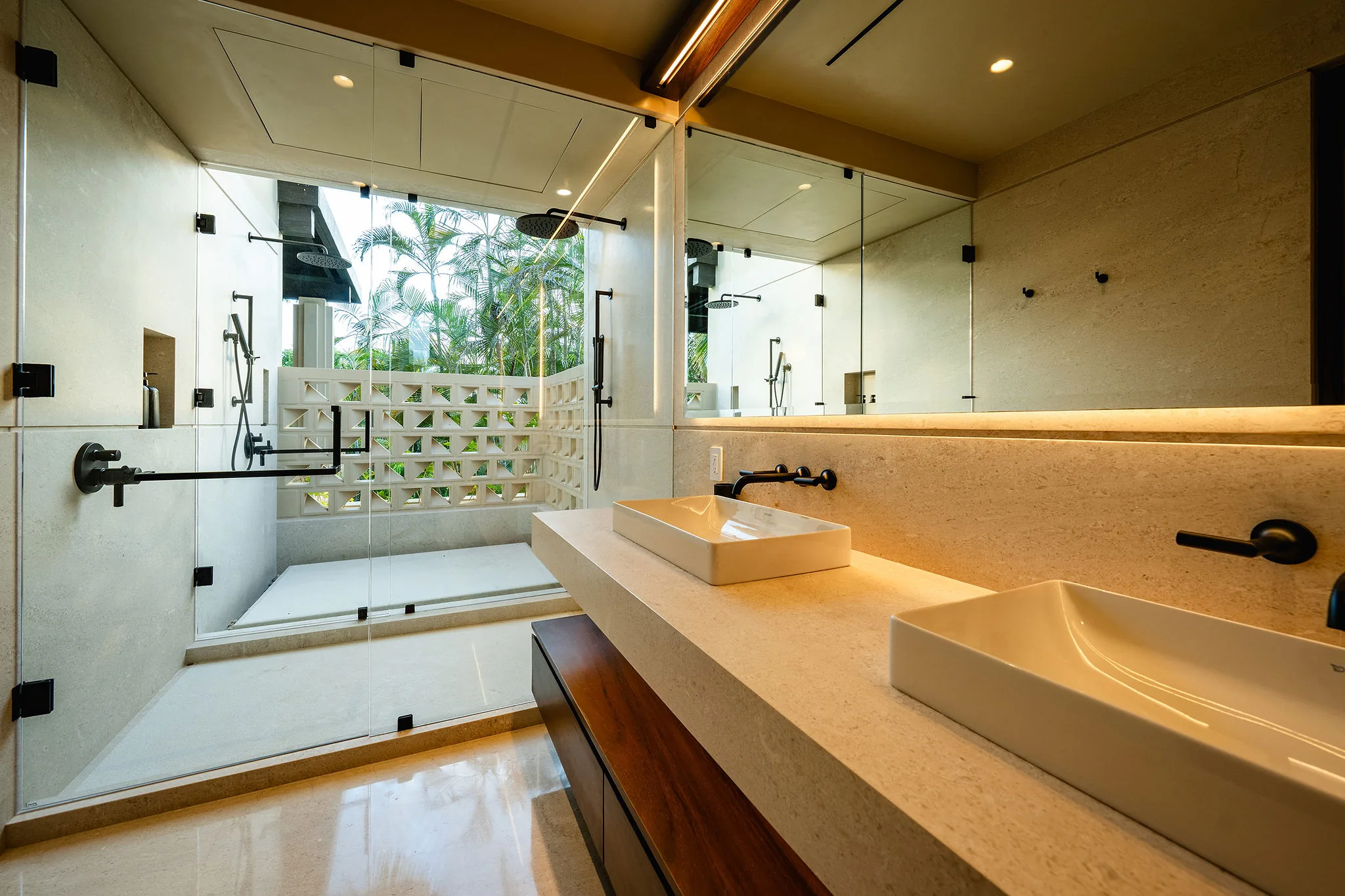 Ensuite bath with double vessel sinks on a stone-and-teak vanity, LED mirror strip, and a rain-head glass shower open to a lattice garden wall with palms, Casa Happy Paws, Ranchos Estates, Punta Mita.