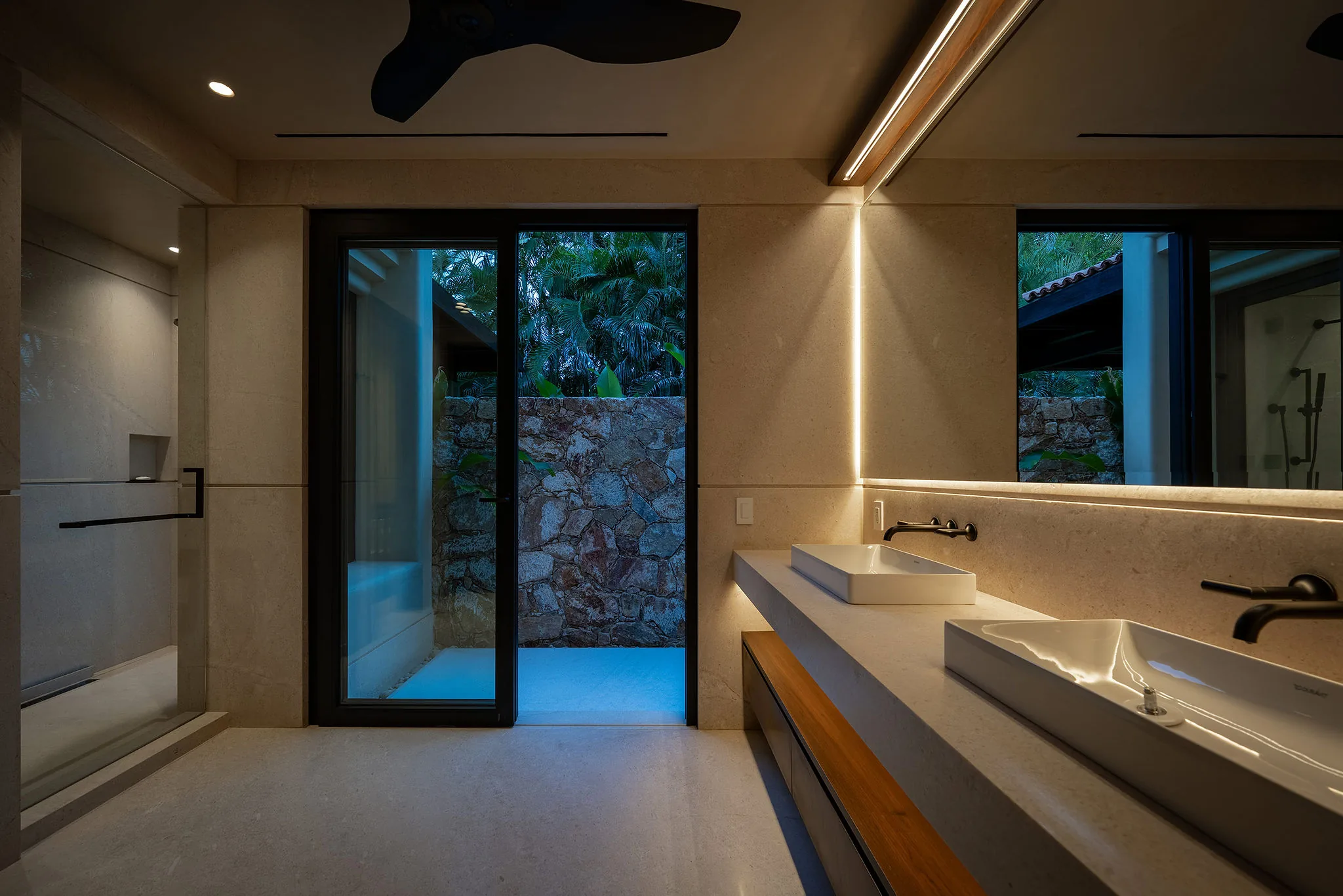 Ensuite bath at dusk with double sinks on a floating stone vanity, LED mirror, and an outdoor shower terrace with stone wall through a sliding glass door, Casa Happy Paws, Ranchos Estates, Punta Mita.