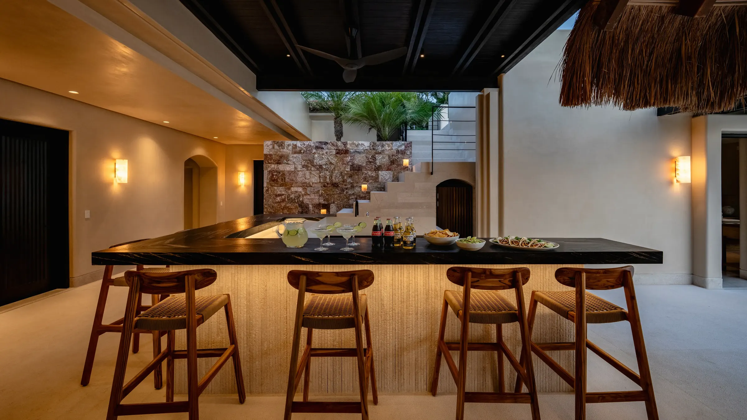 Indoor wet bar with a black marble counter, four cane barstools, margaritas and snacks set out, and an open stone-accent staircase beyond, Casa Happy Paws, Ranchos Estates, Punta Mita.