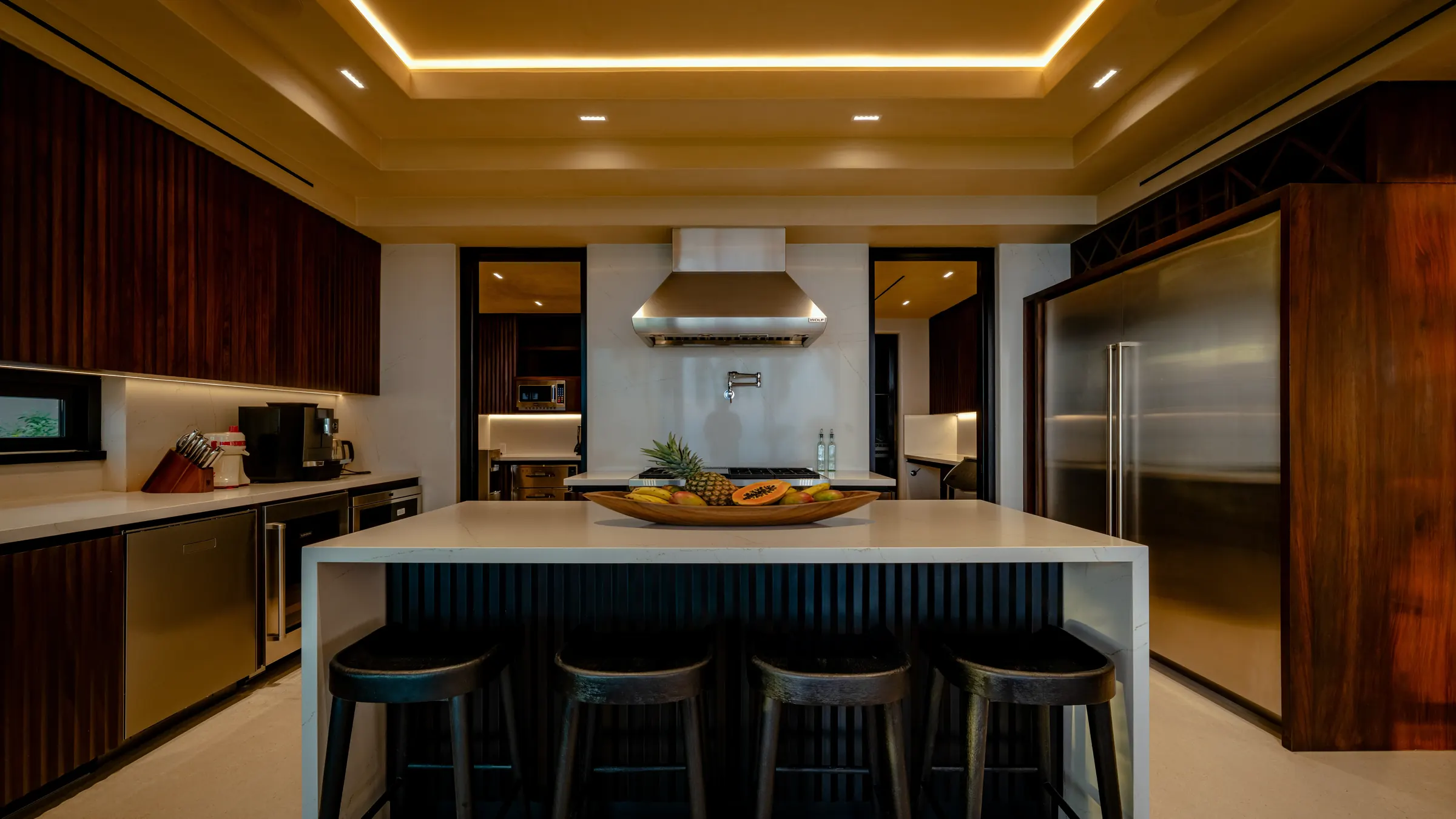 Full chef's kitchen with dark wood ribbed cabinetry, white marble island with four stools, stainless range hood above gas range and a papaya fruit bowl as centerpiece, Casa Happy Paws, Ranchos Estates, Punta Mita.