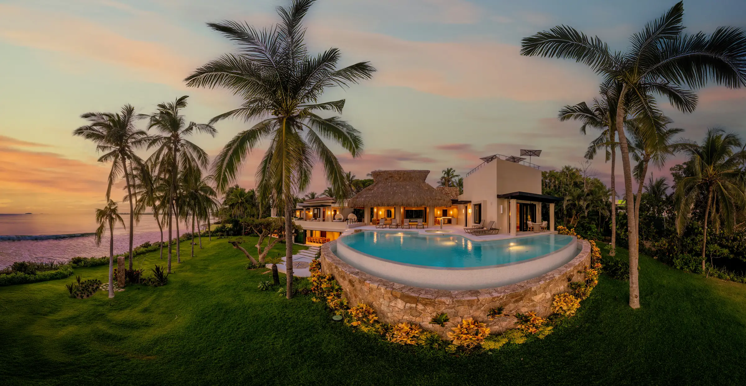 Aerial view at sunset of the crescent pool on its stone plinth surrounded by manicured lawn and palm trees, thatched palapa pavilion and white villa behind, Banderas Bay in the distance, Casa Happy Paws, Ranchos Estates, Punta Mita.