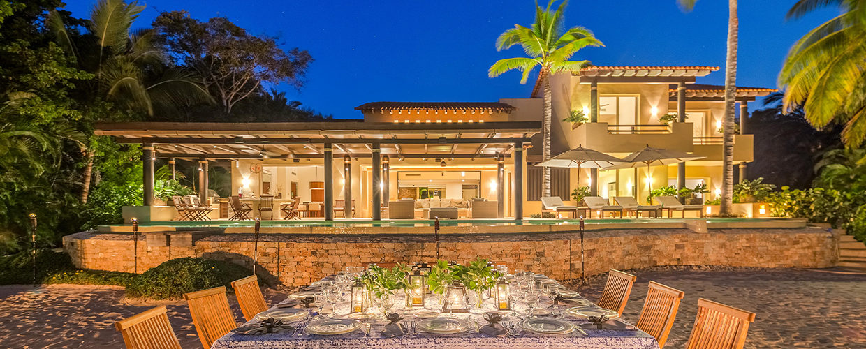 Night exterior from the beach: a wide candlelit dinner table on sand in the foreground, villa fully lit behind with pool terrace above the stone retaining wall, palms and deep blue sky overhead, Casa Manzanillas, Ranchos Estates, Punta Mita.