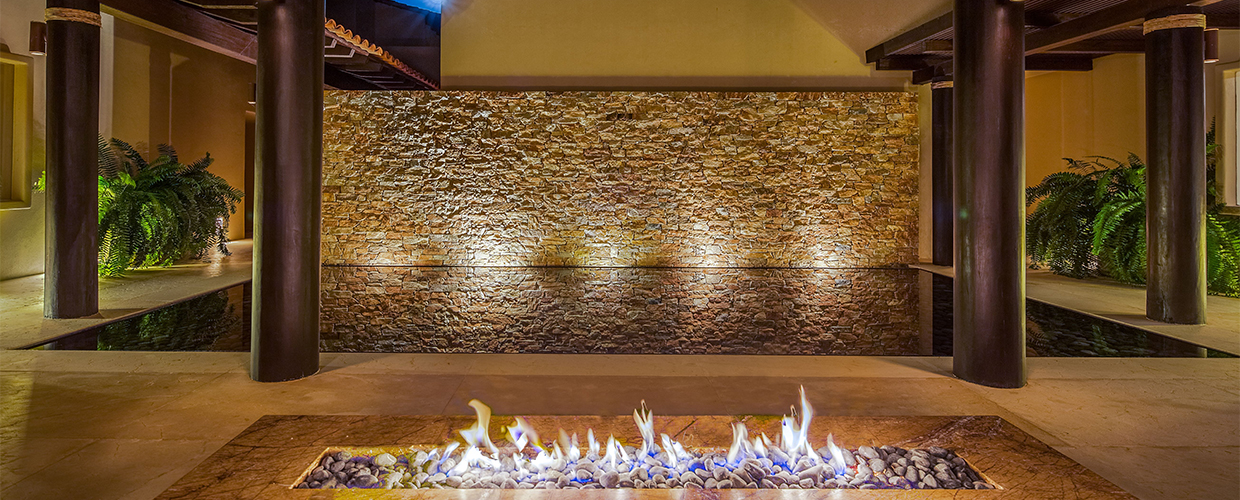Courtyard fire trough at night from a low wide angle: flames run along the stone-pebble channel, rough-cut stone wall glows amber behind, dark timber columns frame each side, ferns lit at the base, Casa Manzanillas, Ranchos Estates, Punta Mita.