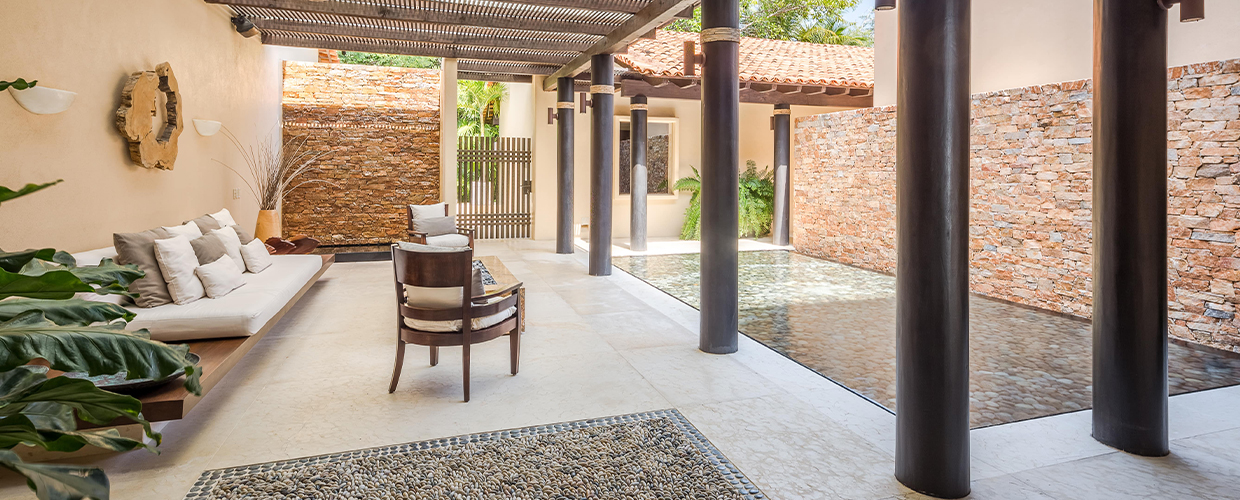 Arrival courtyard in warm daylight: corrugated-steel pergola overhead, dark rope-wrapped columns, built-in daybed with cushions at left, single armchair at centre, pebble mosaic rug, stone-clad walls catching afternoon sun, Casa Manzanillas, Ranchos Estates, Punta Mita.